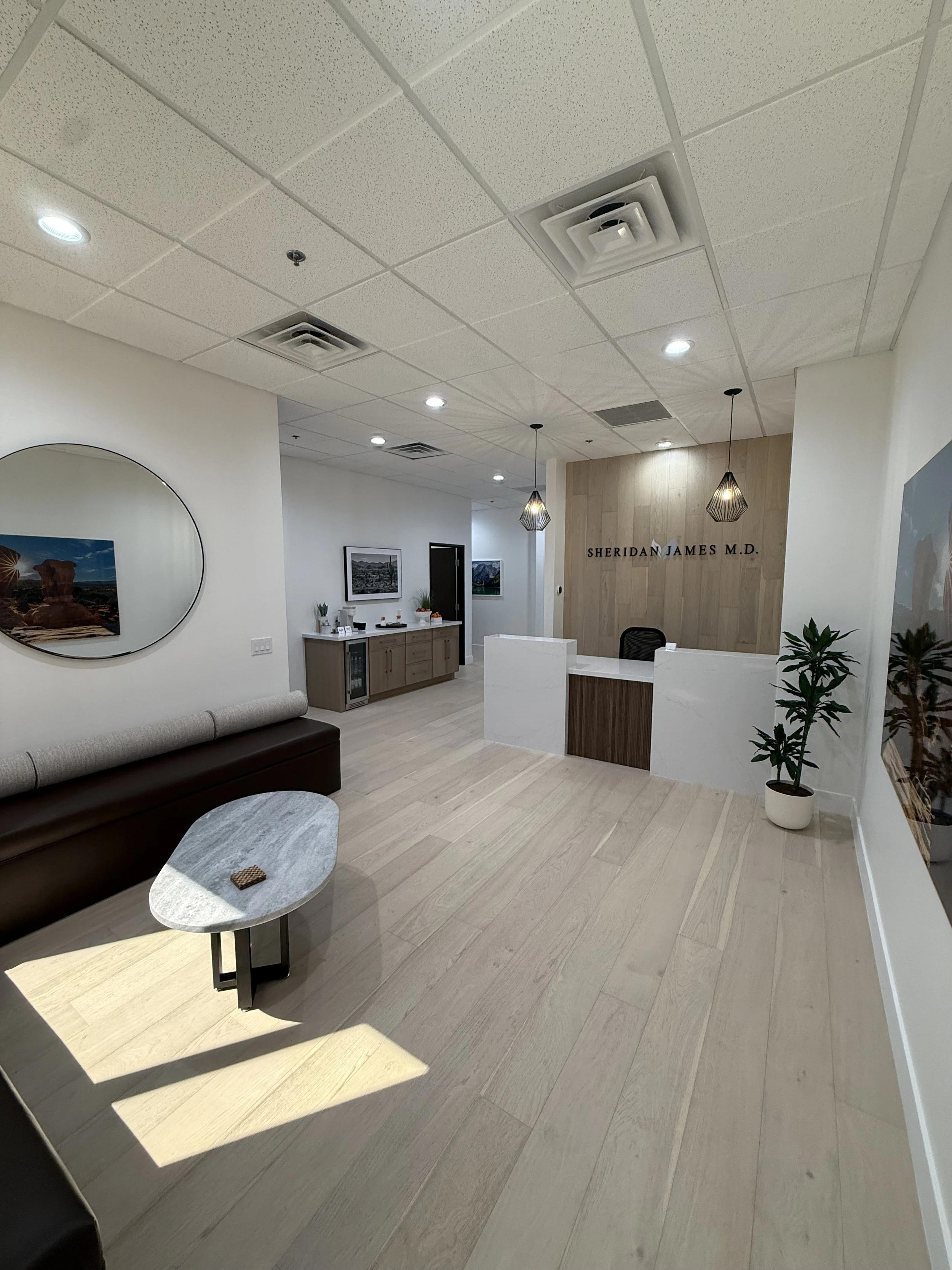Modern medical office reception area with a marble-topped coffee table, brown leather seating, wall art, potted plant, and a reception desk with the name 'Sheridan James M.D.' on the wall behind.