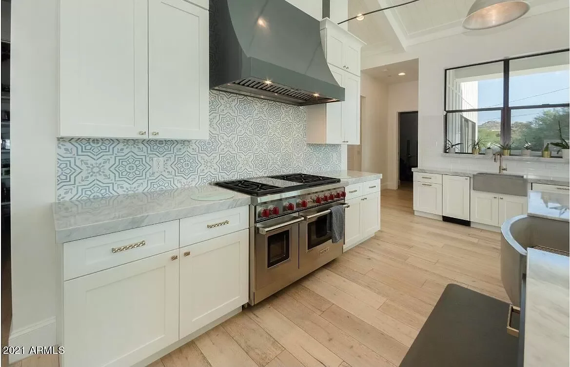 Modern kitchen with white cabinets, a patterned tile backsplash, a large stainless steel stove with red knobs, a stainless steel range hood, a window above the sink, and light wooden flooring.