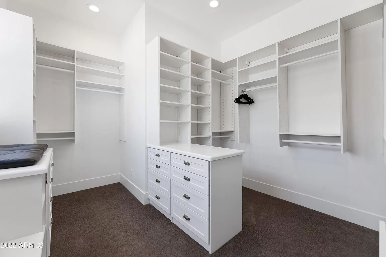 Empty walk-in closet with white built-in shelves, drawers, and hanging rods. Brown carpeted floor and white walls.