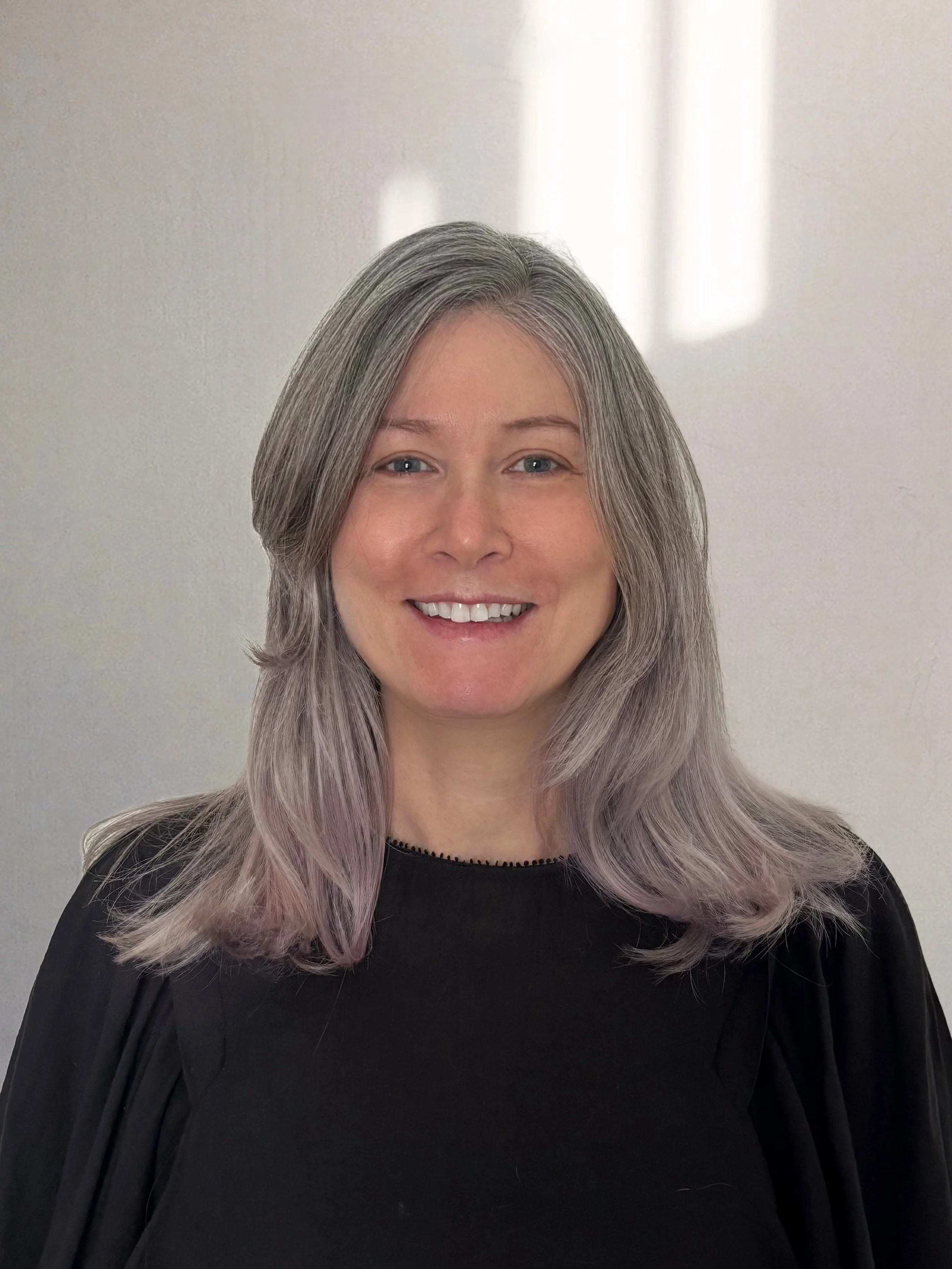 Portrait of a smiling woman with grayish-lavender hair, wearing a black top, standing against a plain background with soft light.