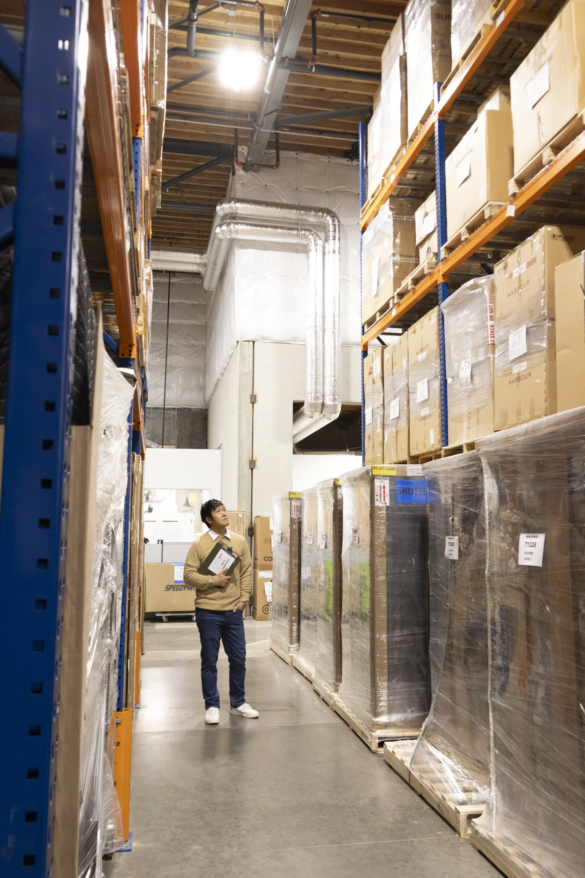 A person standing in a warehouse aisle among stacked pallets and boxes, with industrial shelving and ductwork overhead.