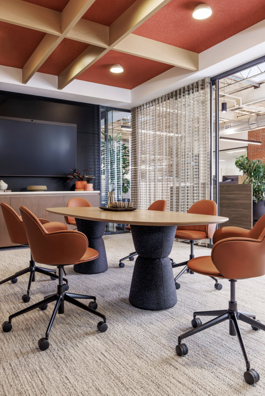 Modern conference room with round wooden table, six orange office chairs with wheels, a large flat-screen TV on the wall, a black cabinet with decorative items, plants, and a glass wall with patterned curtains. Open ceiling with exposed ducts and a mix of natural and artificial lighting.