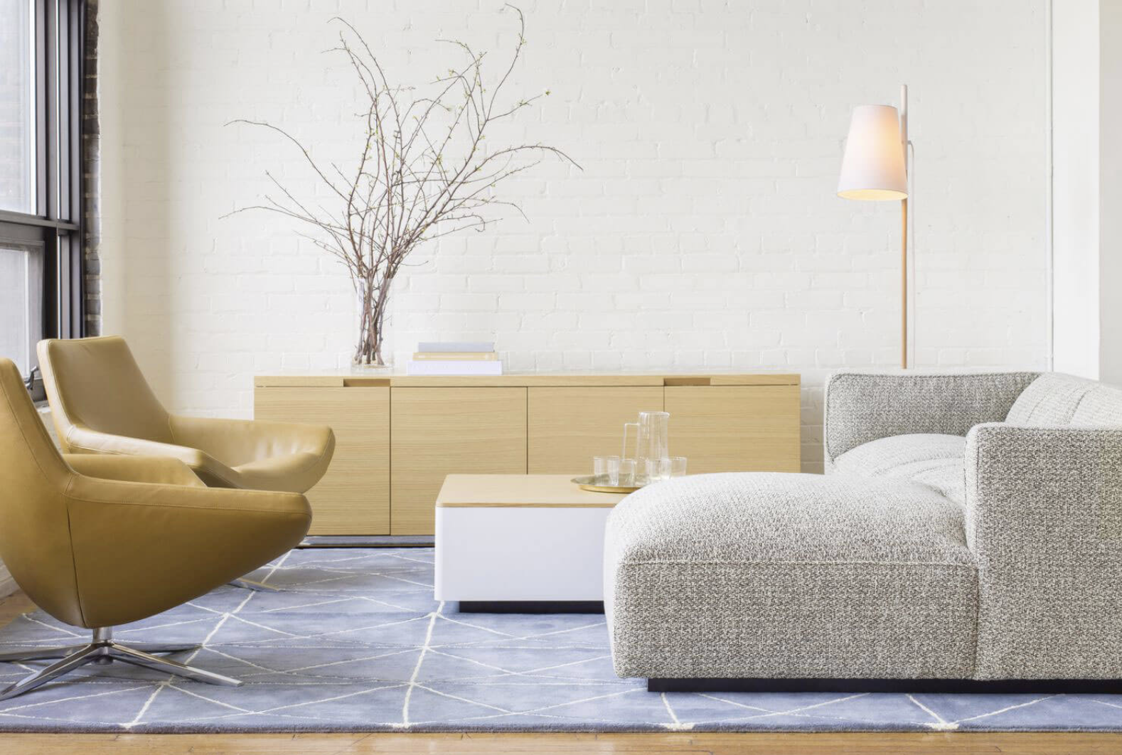 Modern living room with a gray sofa, gold chairs, a wooden cabinet, a side table with glasses, floor lamp, and bare branches in a vase near a window.