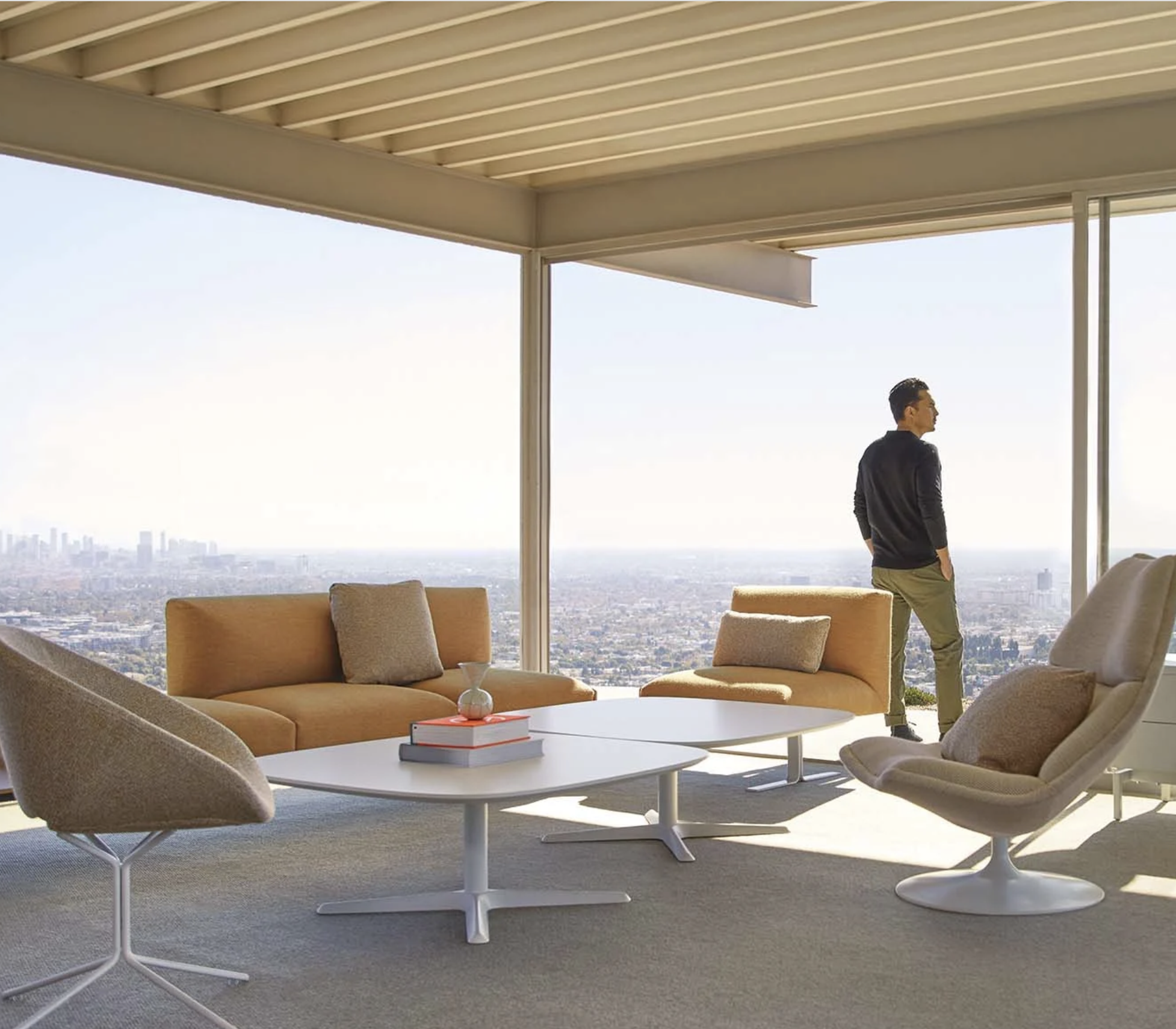 A modern interior living room with large floor-to-ceiling windows overlooking a cityscape. The room has beige and light brown furniture, including a sofa, armchairs, and a coffee table with books and a small decorative item. A man stands near the win
