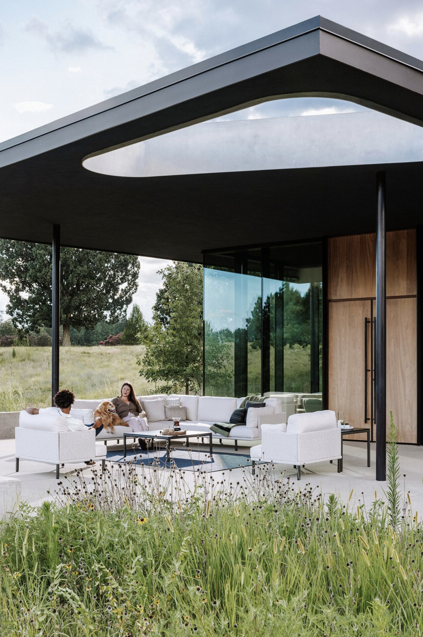 Modern outdoor patio with white furniture, a woman and a man sitting with a dog, surrounded by greenery and trees, with a contemporary house featuring a large glass window and a wooden wall.