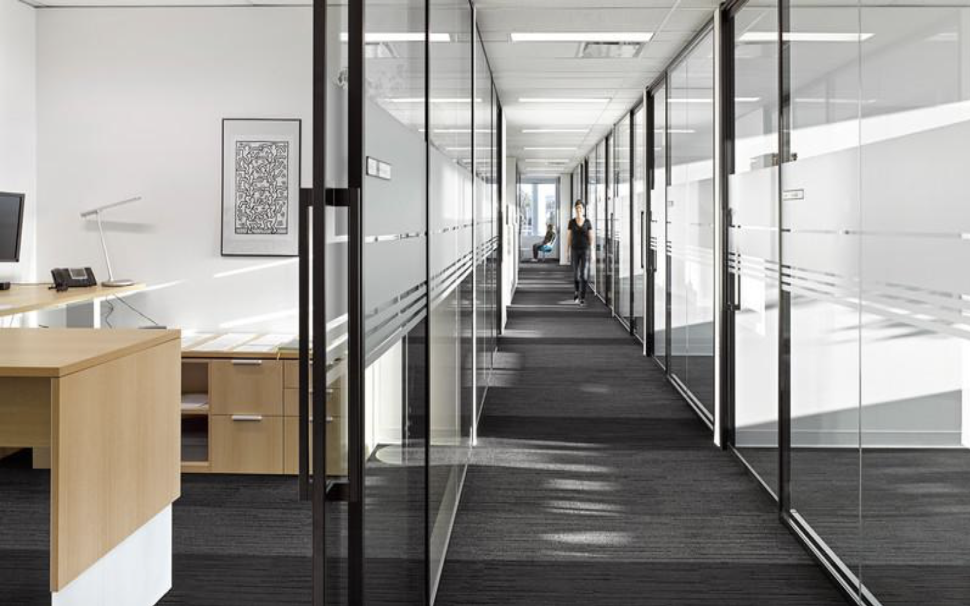 Modern office hallway with glass walls and doors, black carpet, and a person walking toward the camera.