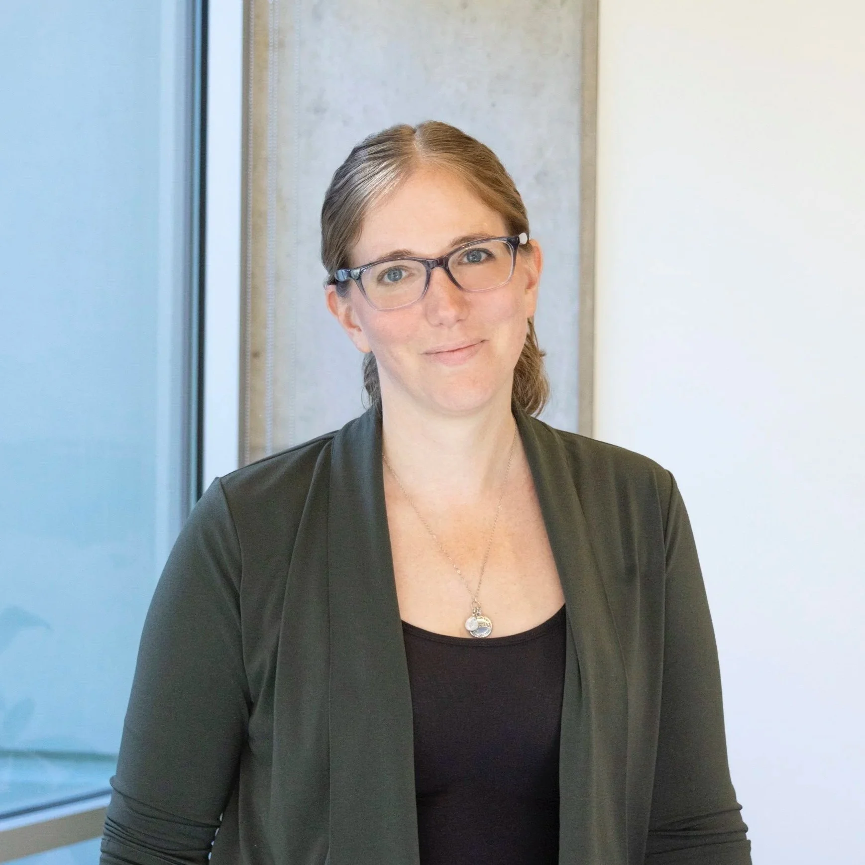 A woman with light skin, brown hair, glasses, wearing a black top and a dark green blazer with a silver necklace, standing indoors near a window with a neutral expression.
