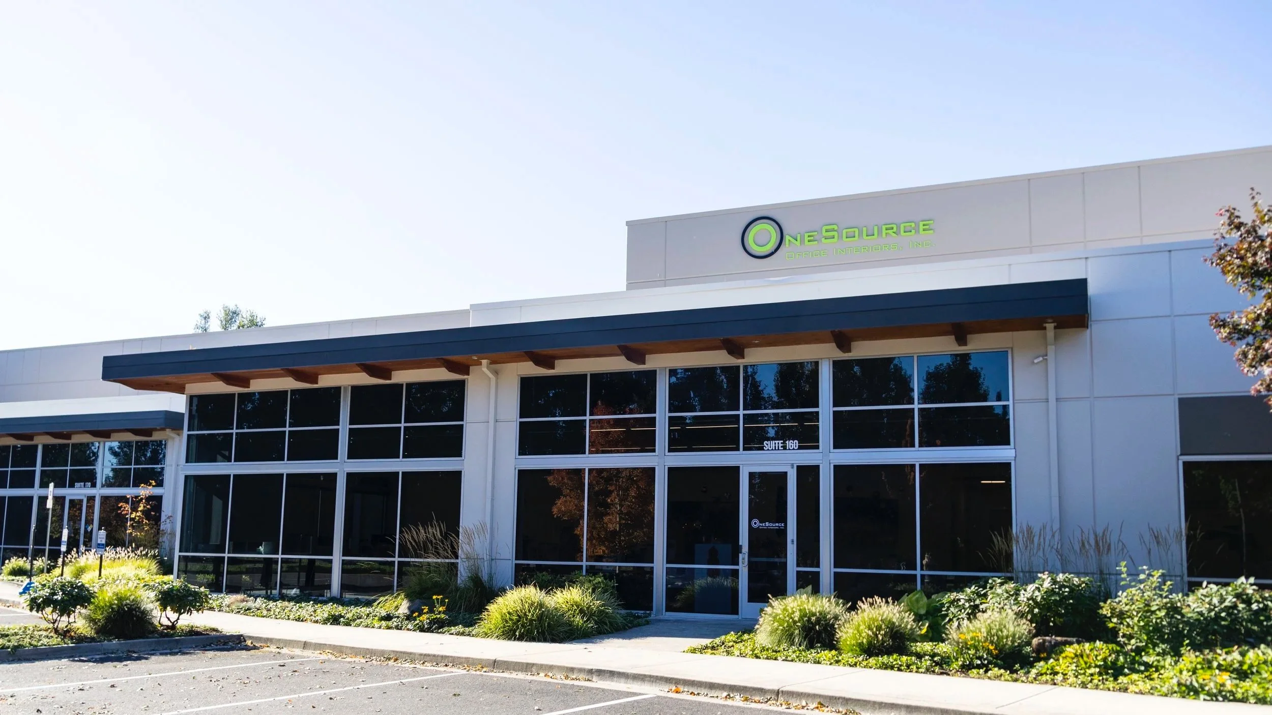 Front view of a modern office building with large glass windows, a sign reading 'OneSource Office Interiors Inc.', landscaped bushes, and a parking lot.