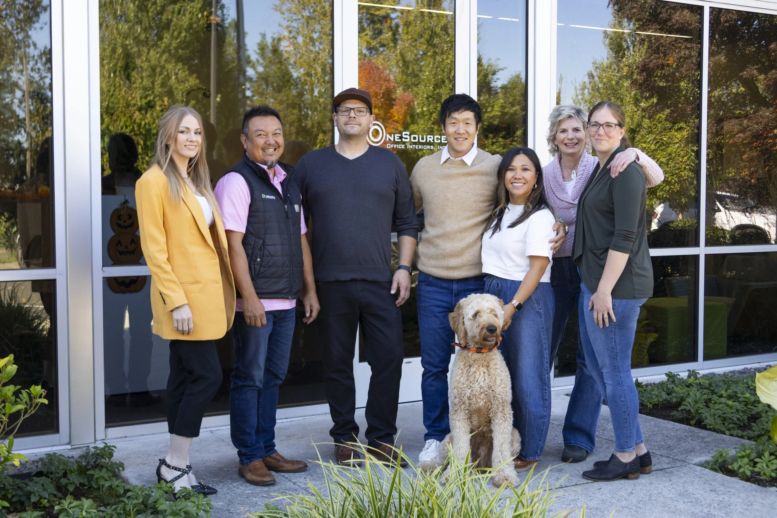 Group of eight people standing outside a building with large glass windows, smiling, with a dog sitting in front of them. The reflection in the windows shows trees and a parking lot. The building has a sign that reads "OneSource Office Interiors, Inc."