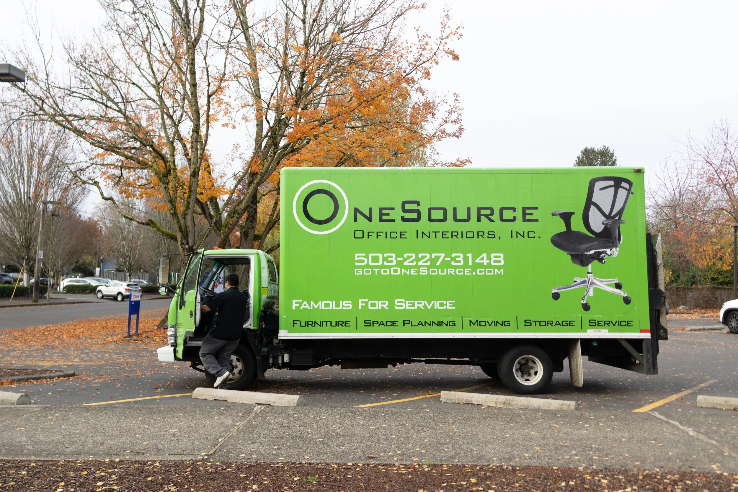 A green moving truck is parked in a parking lot with autumn leaves on the ground and trees with orange and yellow foliage in the background. A person is stepping into the truck's cab, and the truck has advertisements for NeSource Office Interiors, Inc., including their phone number and website, and services like furniture, space planning, moving, storage, and service.