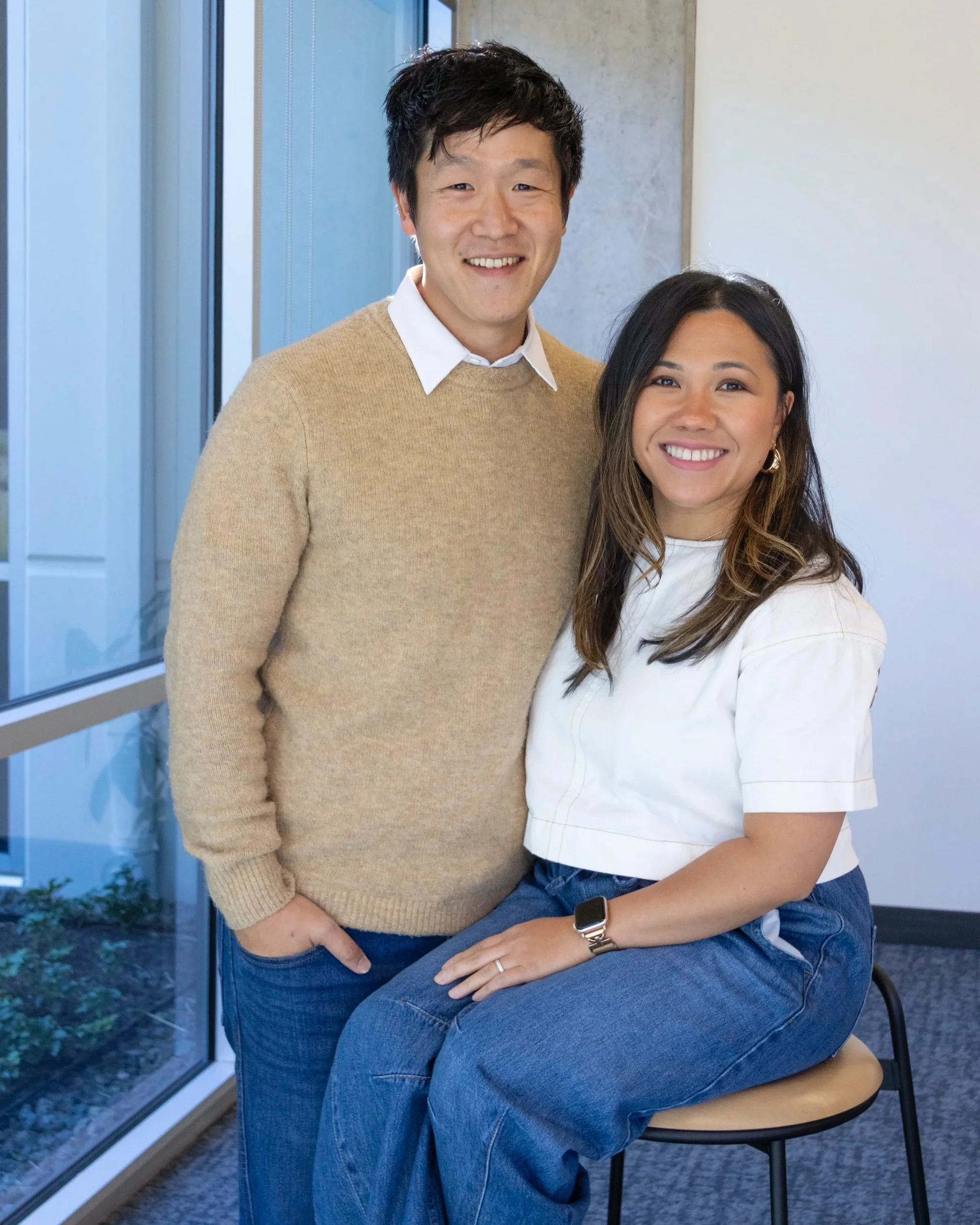 A man and a woman smiling for a photo indoors near a window, with the woman sitting on a chair.