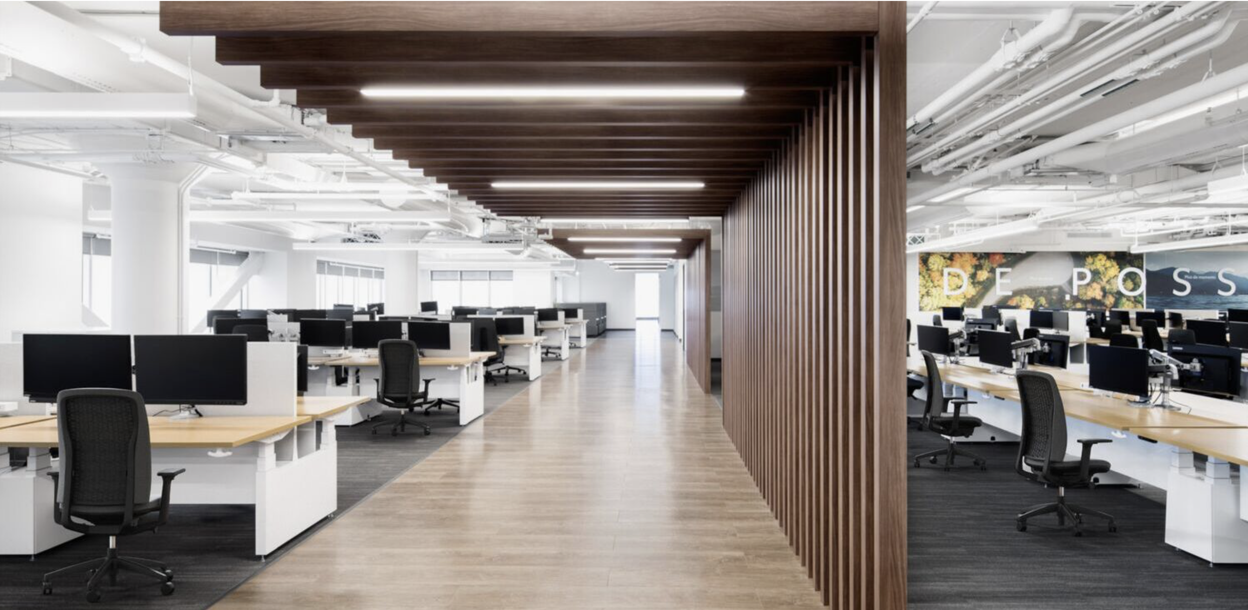 Modern open-plan office with rows of desks and chairs, divided by a wooden slatted partition, bright lighting, and large windows.