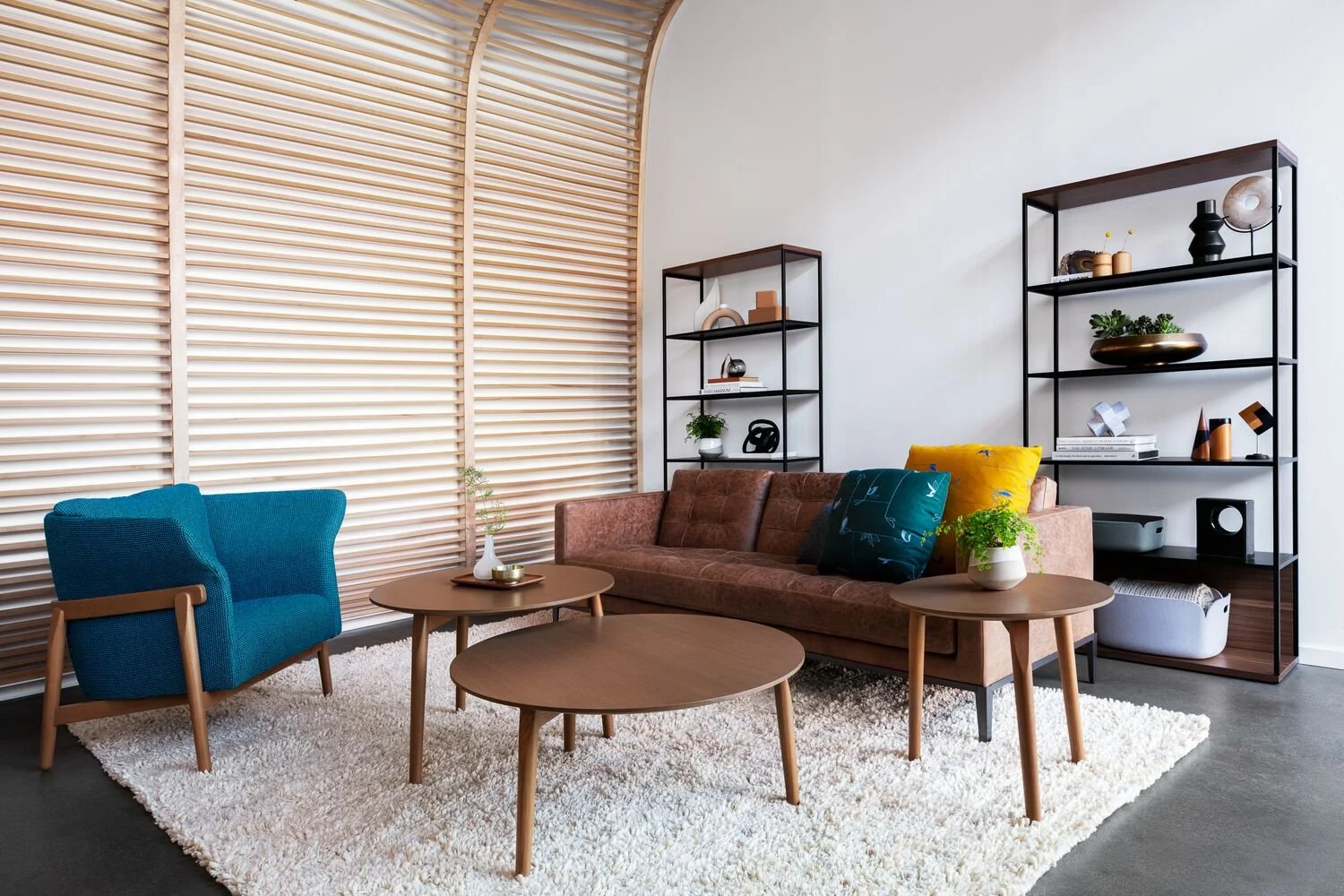 Modern living room with a brown sofa, blue armchair, three round wooden coffee tables, and black metal shelves with decorative items. Light wood blinds cover the large window, and a white shag rug covers the dark floor.