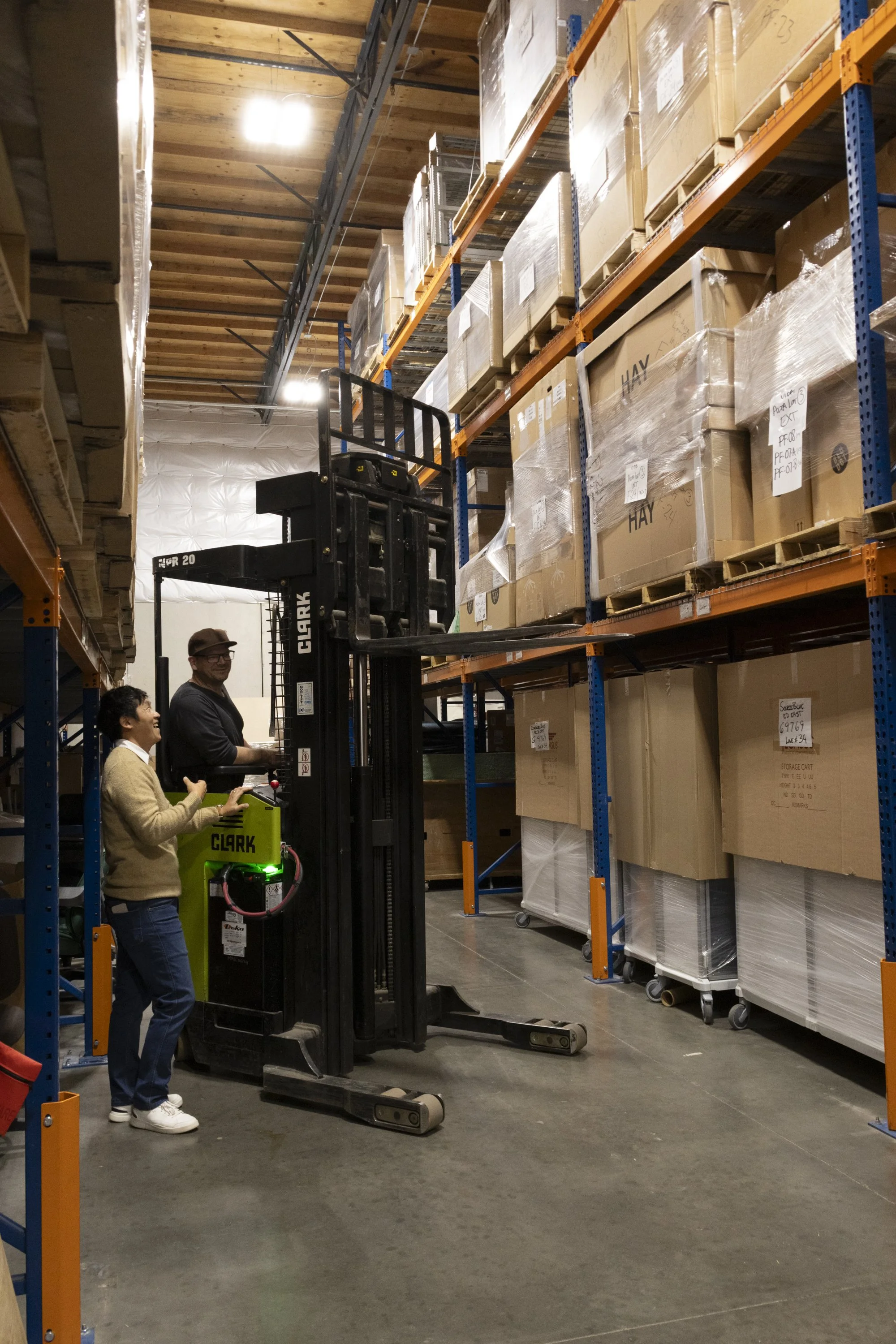 Two people operating a forklift in a warehouse aisle filled with stacked boxes on industrial shelving.