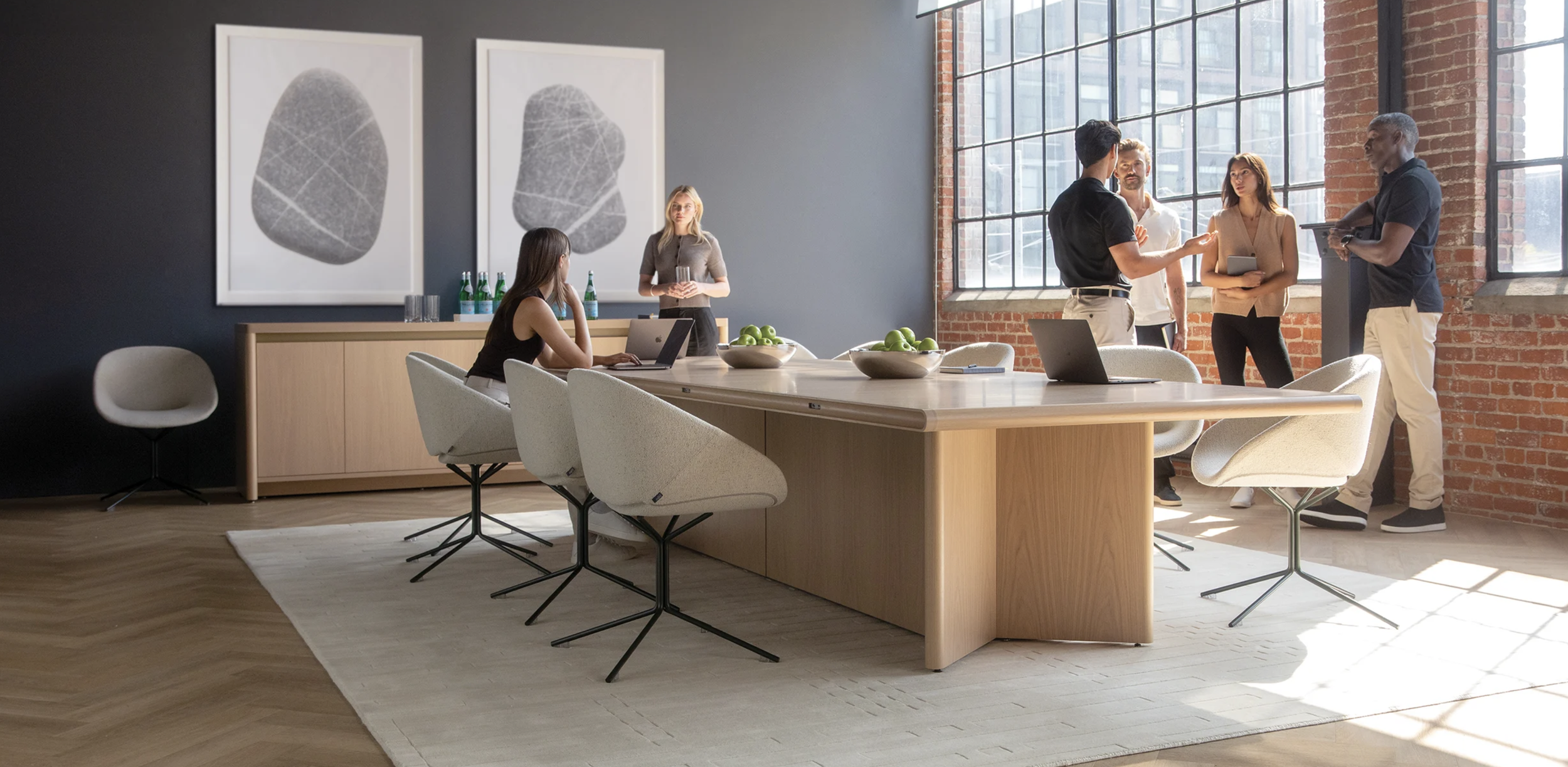 A modern office meeting room with five people, a large wooden table, laptops, green apples in bowls, and large windows with brick walls.