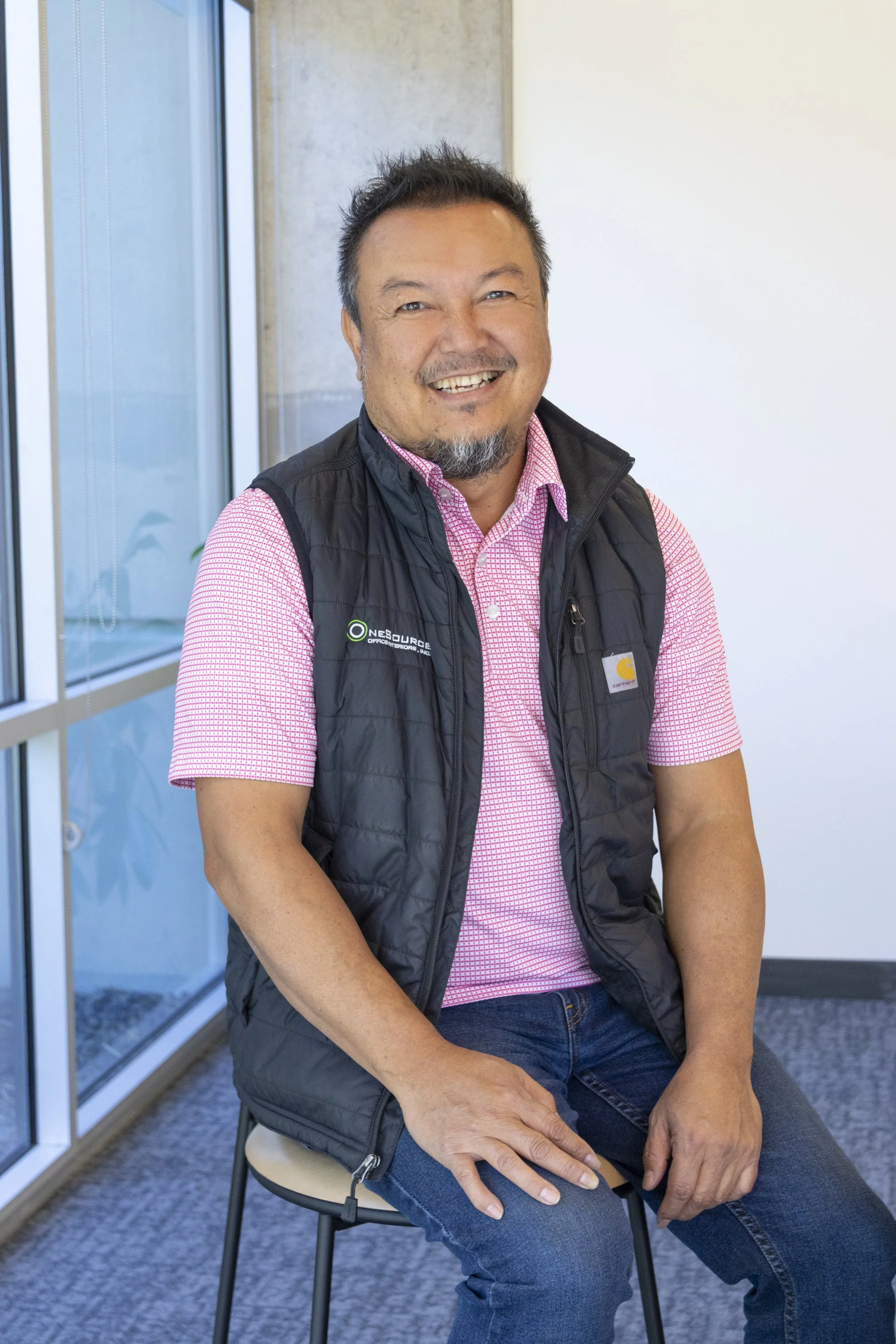 Smiling man with dark hair, beard, and mustache, wearing a pink checkered shirt and black vest, sitting on a beige stool in an office, next to a glass window.