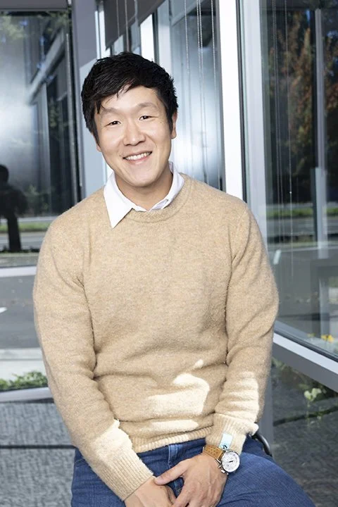 A smiling young man in a beige sweater and white collared shirt sitting by a large window.