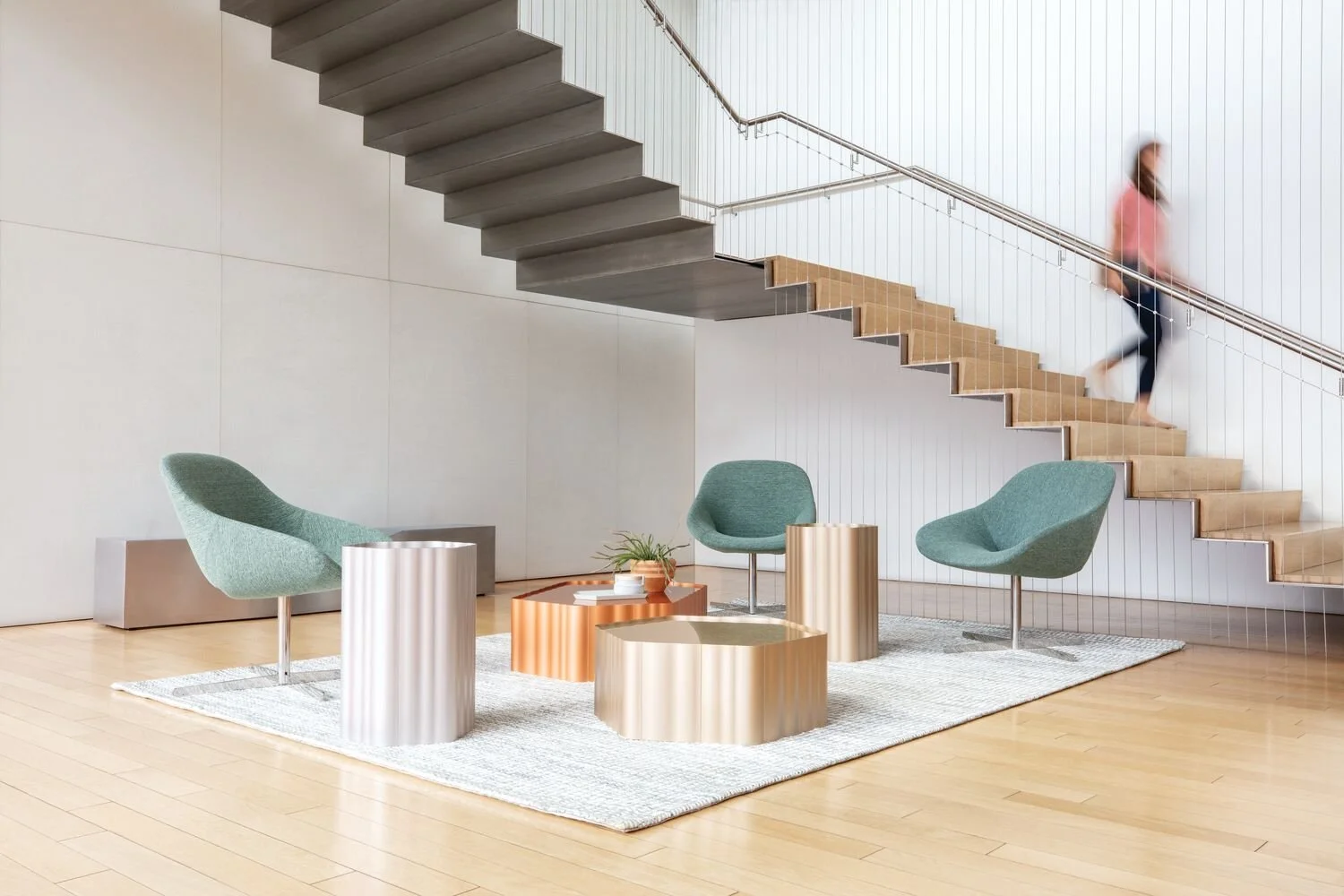 Modern interior with green chairs, metallic side tables, a white rug, a wooden staircase, and a person walking up the stairs.