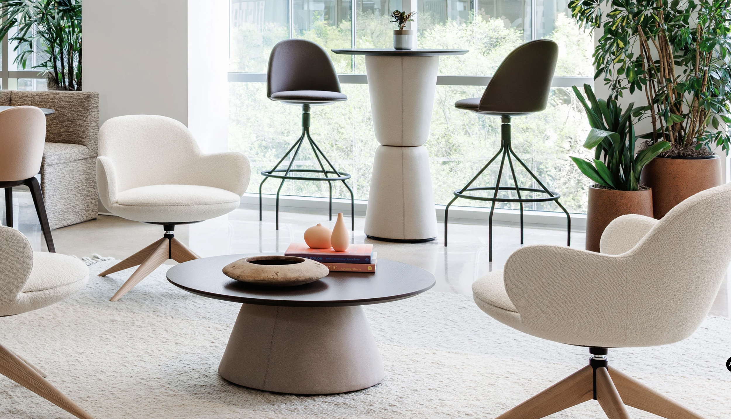 Modern living room with beige and white furniture, a dark wood coffee table with decorative objects, indoor plants, and floor-to-ceiling windows with outdoor greenery.