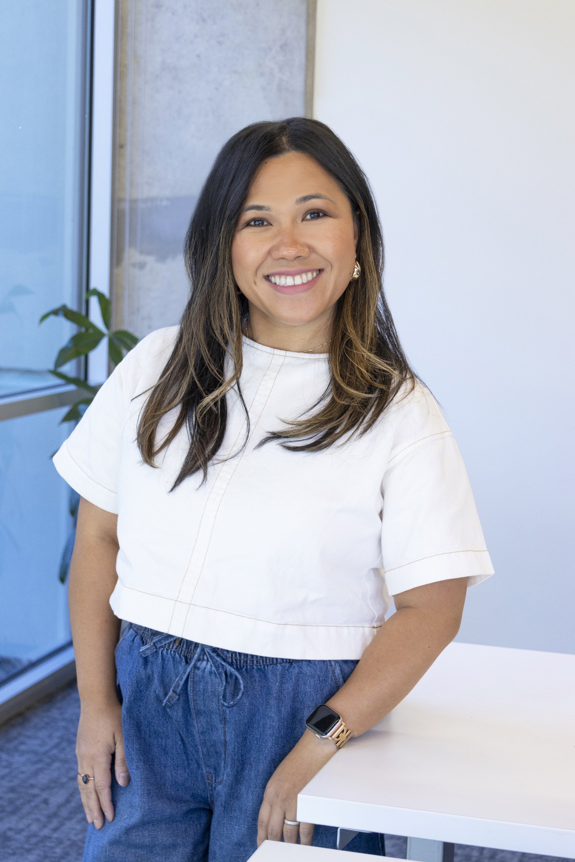 Smiling woman wearing a white top, blue jeans, gold watch, and jewelry, standing beside a white table in an office space with a plant nearby.