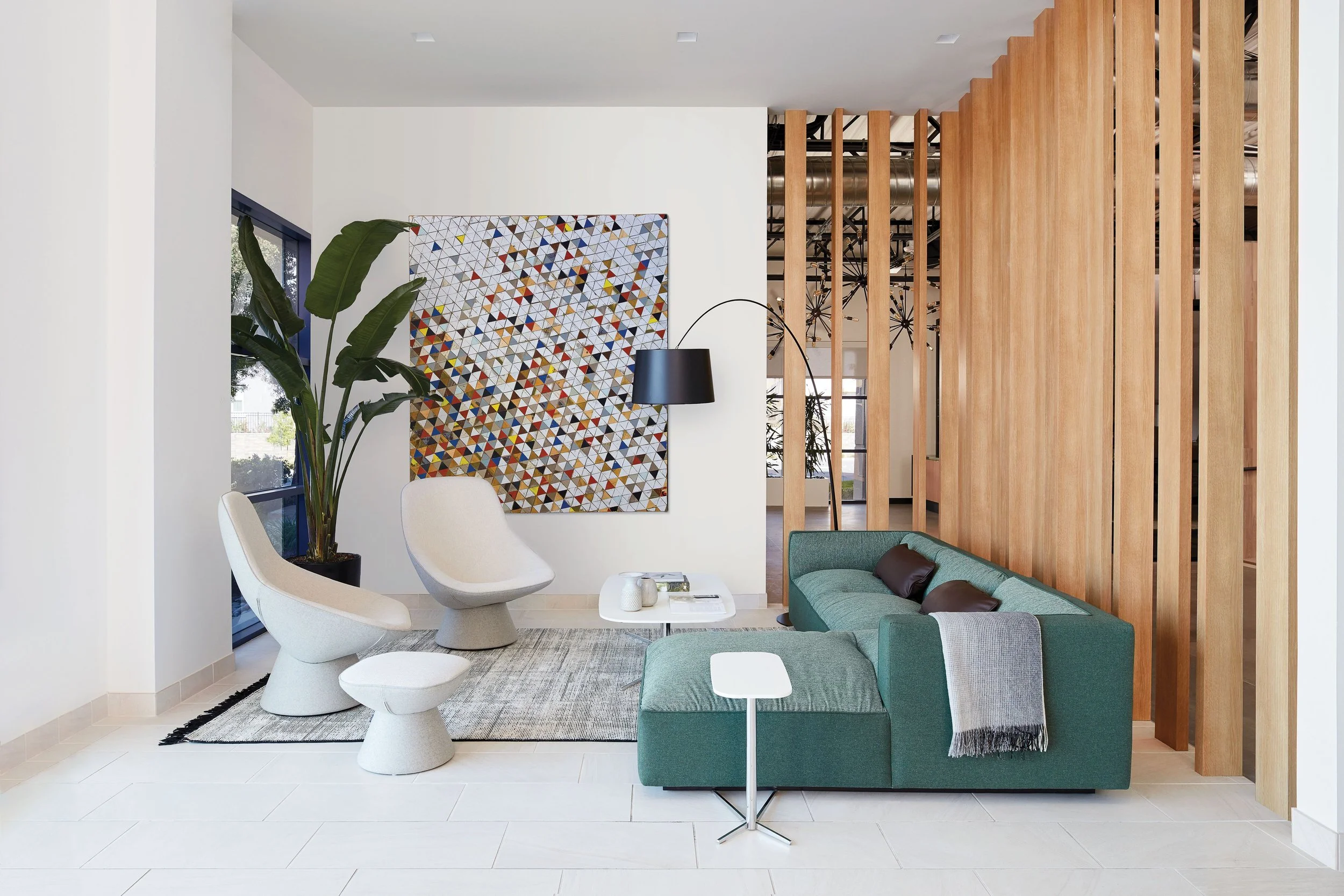 Modern living room with white walls, a green sectional sofa, two white sculptural chairs, a large plant, artwork with a colorful geometric pattern, a black floor lamp, and a wooden slat room divider.