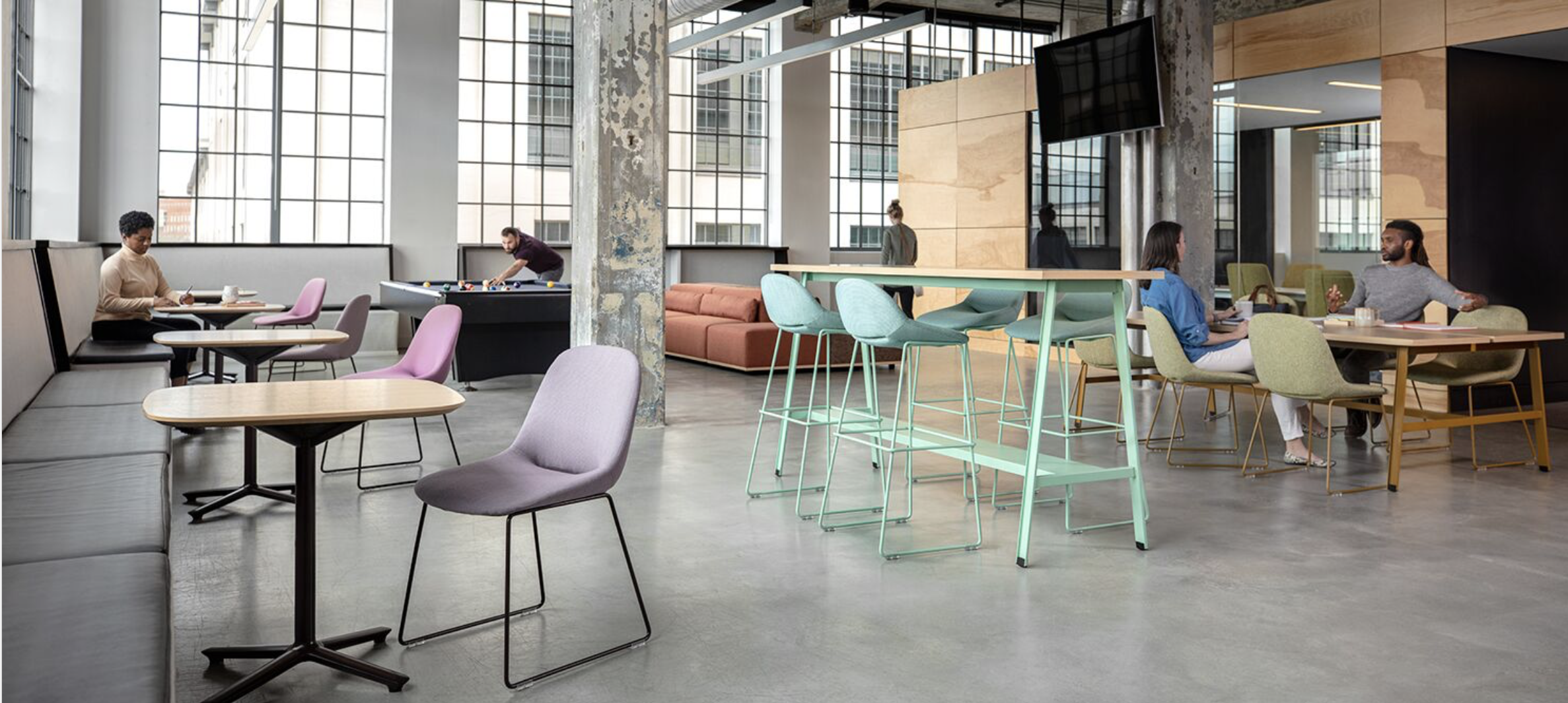 Modern coworking space with people working and playing, including a woman at a table, a man playing pool, and two people talking at a high table, with large windows and mixed seating.