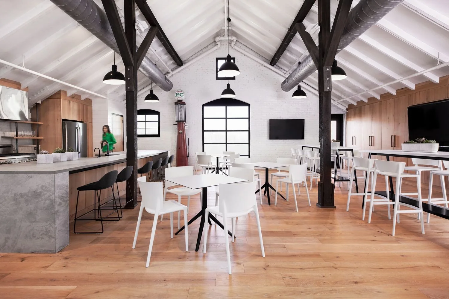 Modern, spacious indoor dining area with high vaulted ceiling, black and white furniture, and wooden flooring. A woman in green is near the kitchen area with bar stools. The space features metal beams, pendant lighting, a wall-mounted TV, and large w