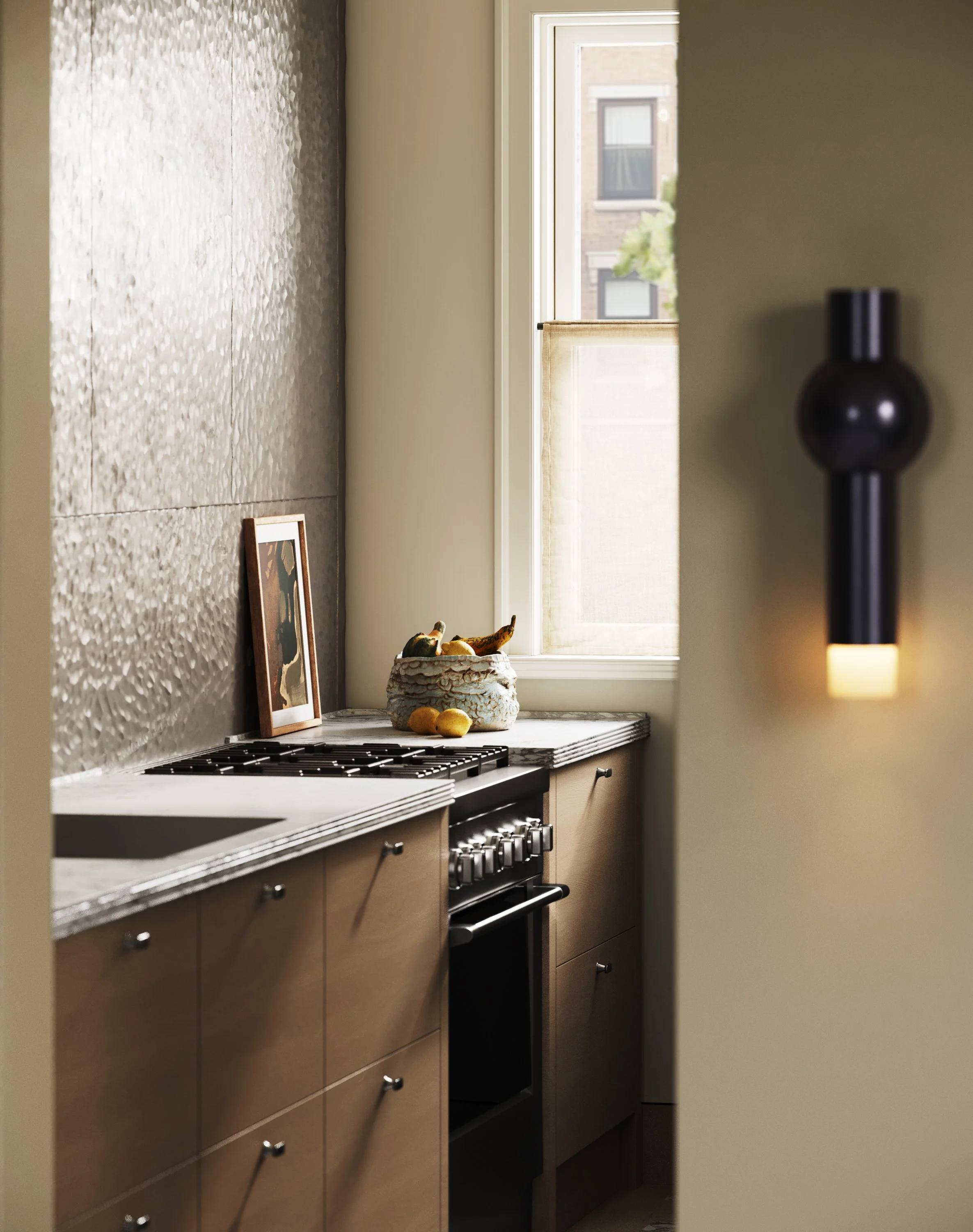 A kitchen with beige cabinets and a marble countertop, a window with sunlight coming through, a framed photo, a bowl with plants, and a black hanging light.