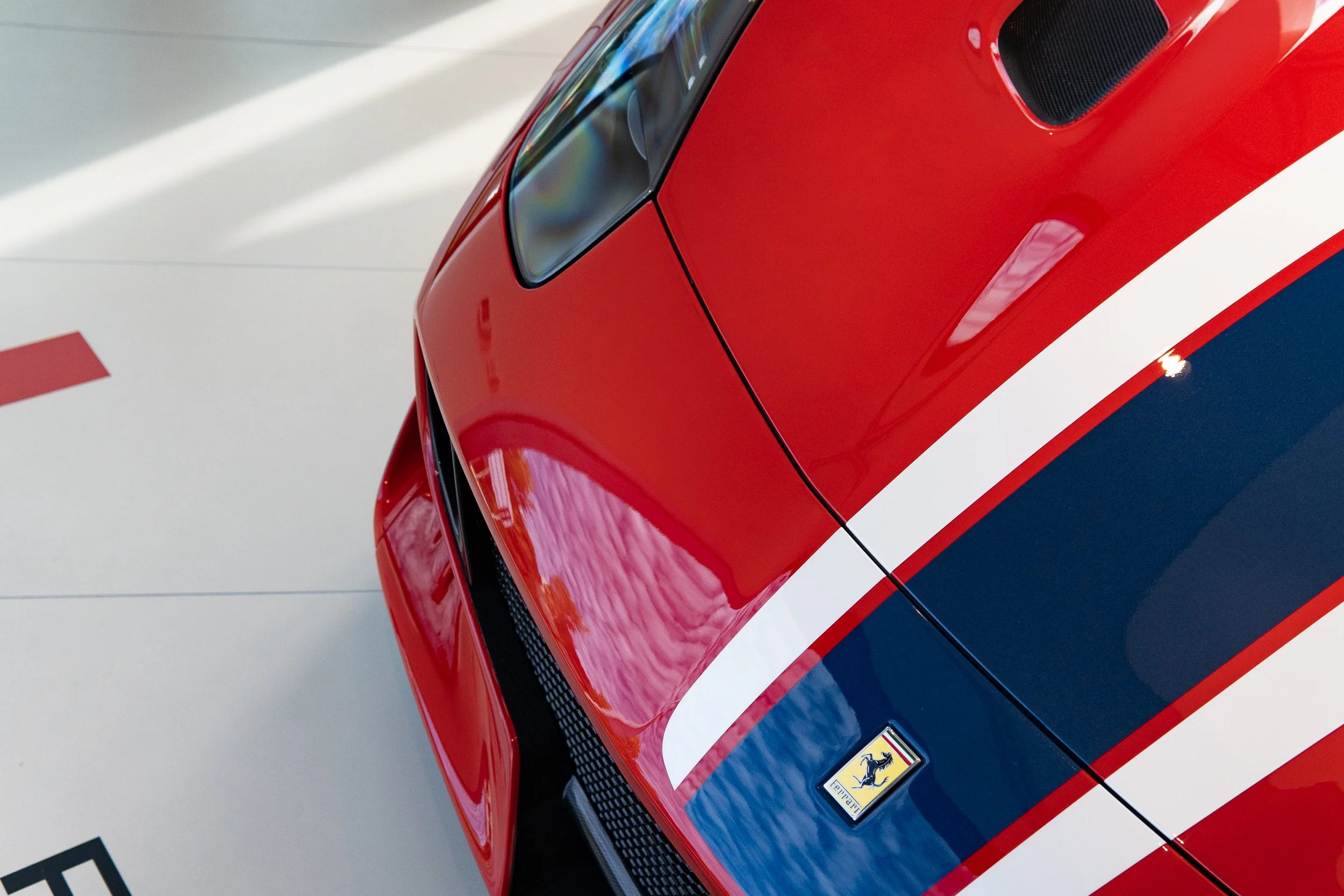 Close-up of the front corner of a red Ferrari sports car with racing stripes and Ferrari emblem.