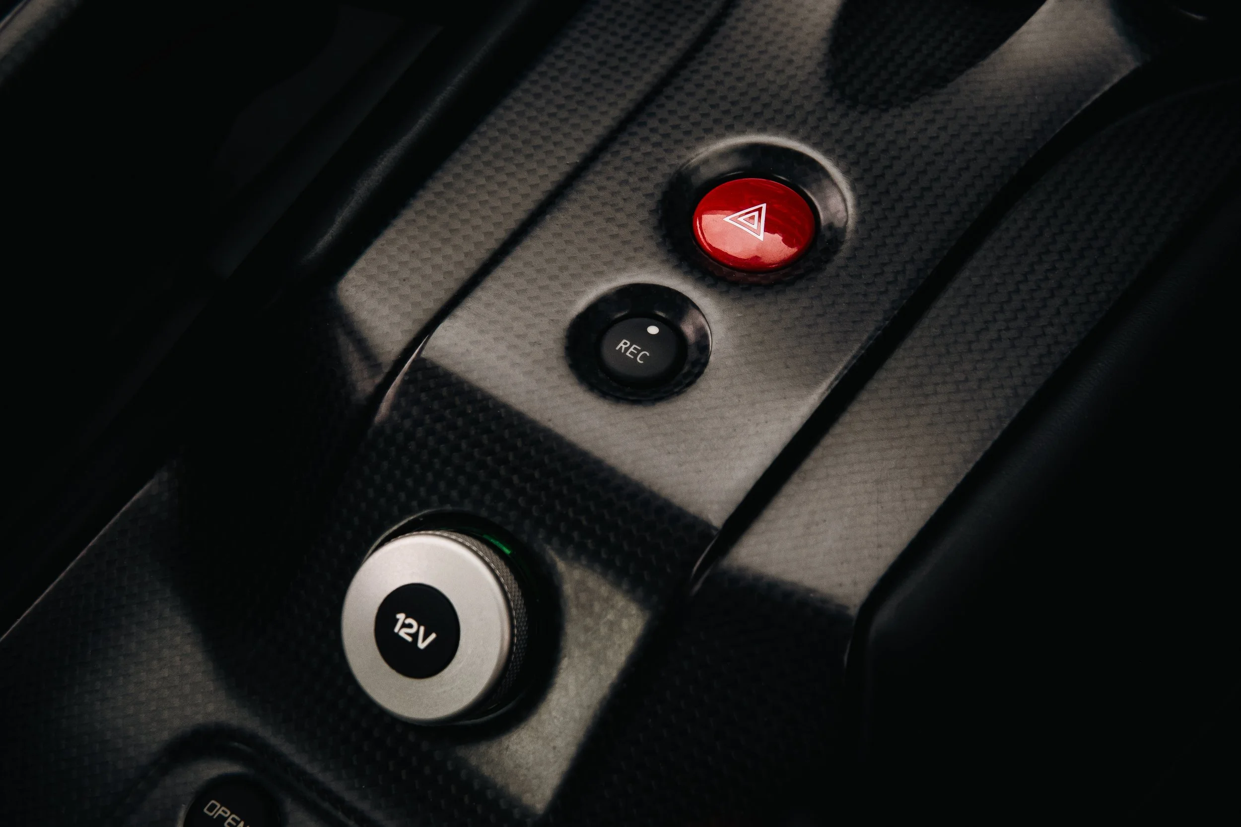 Close-up of a car's dashboard with a red hazard light button, a black 'REC' button, a 12V power outlet, and part of a control panel with textured black surface.