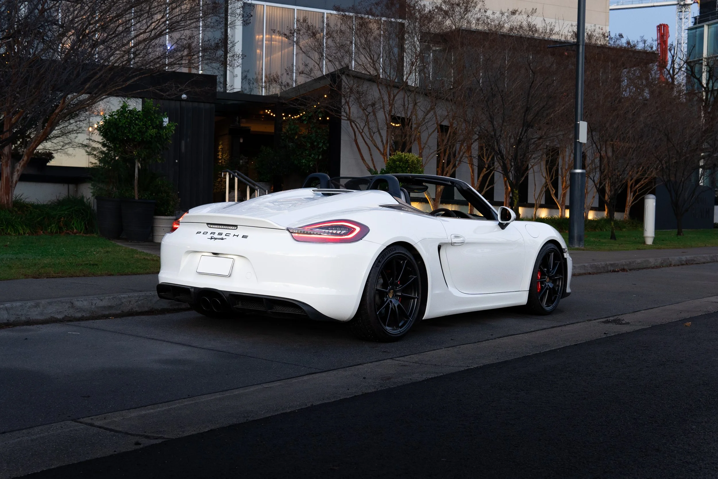 A white Porsche sports car parked on the side of the street in front of a modern building with trees and plants around.