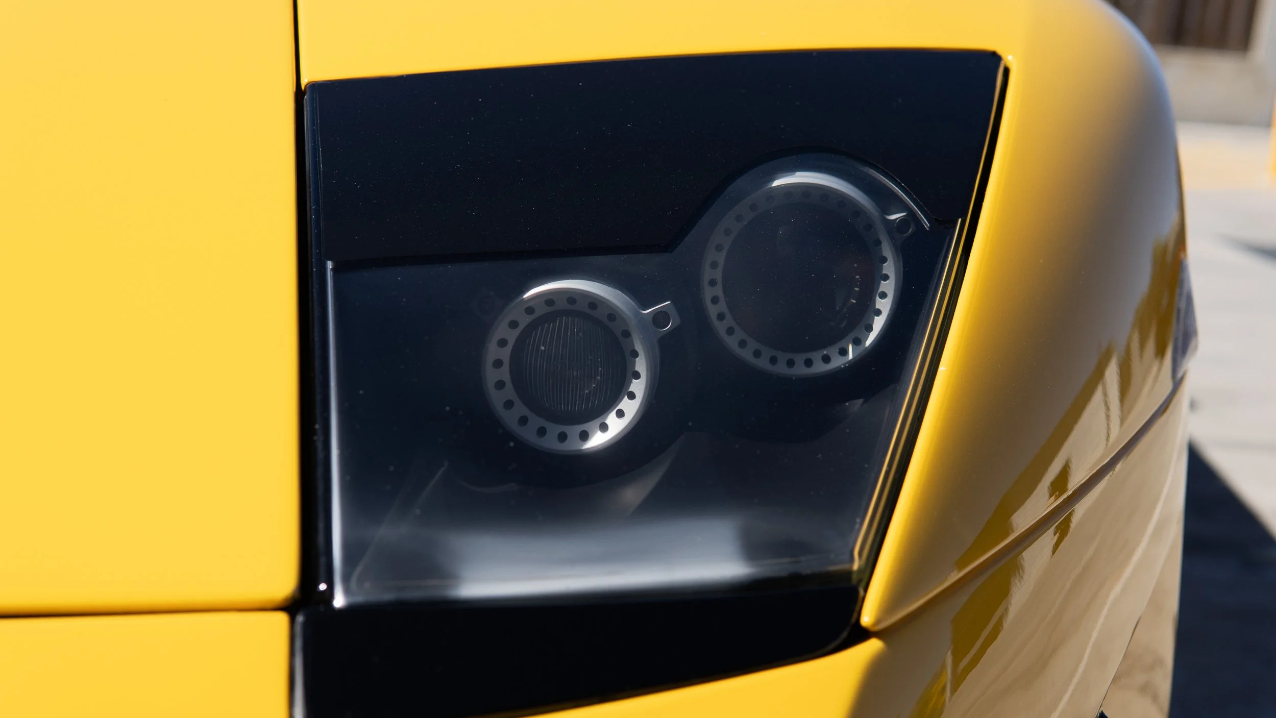 Close-up of a black car headlight with a yellow body in the background.
