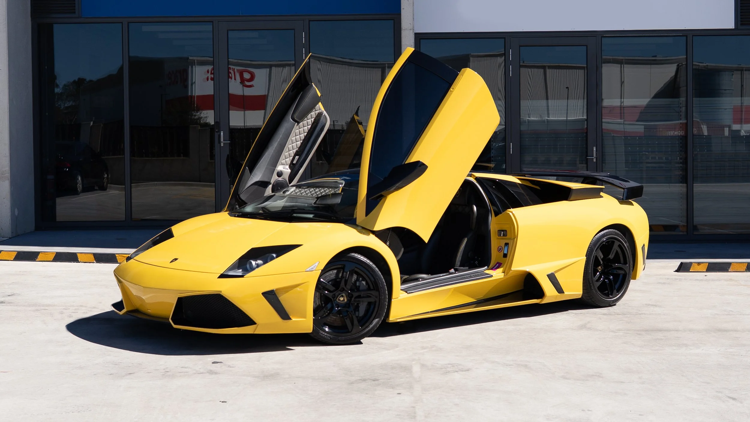 Yellow Lamborghini sports car with scissor doors open, parked outside a modern building with reflective glass windows.