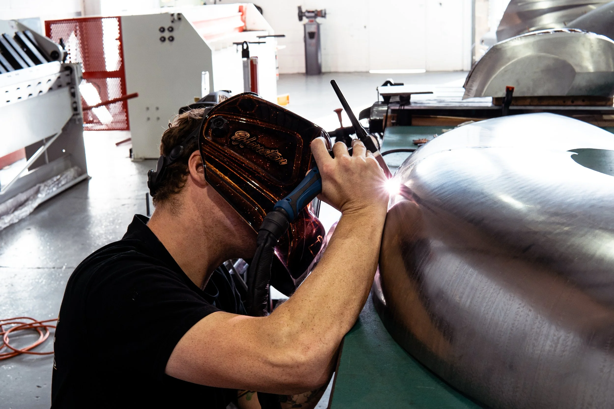A person welding a large metal component in a workshop, wearing a protective welding helmet and gloves, with sparks and bright light from the welding process.