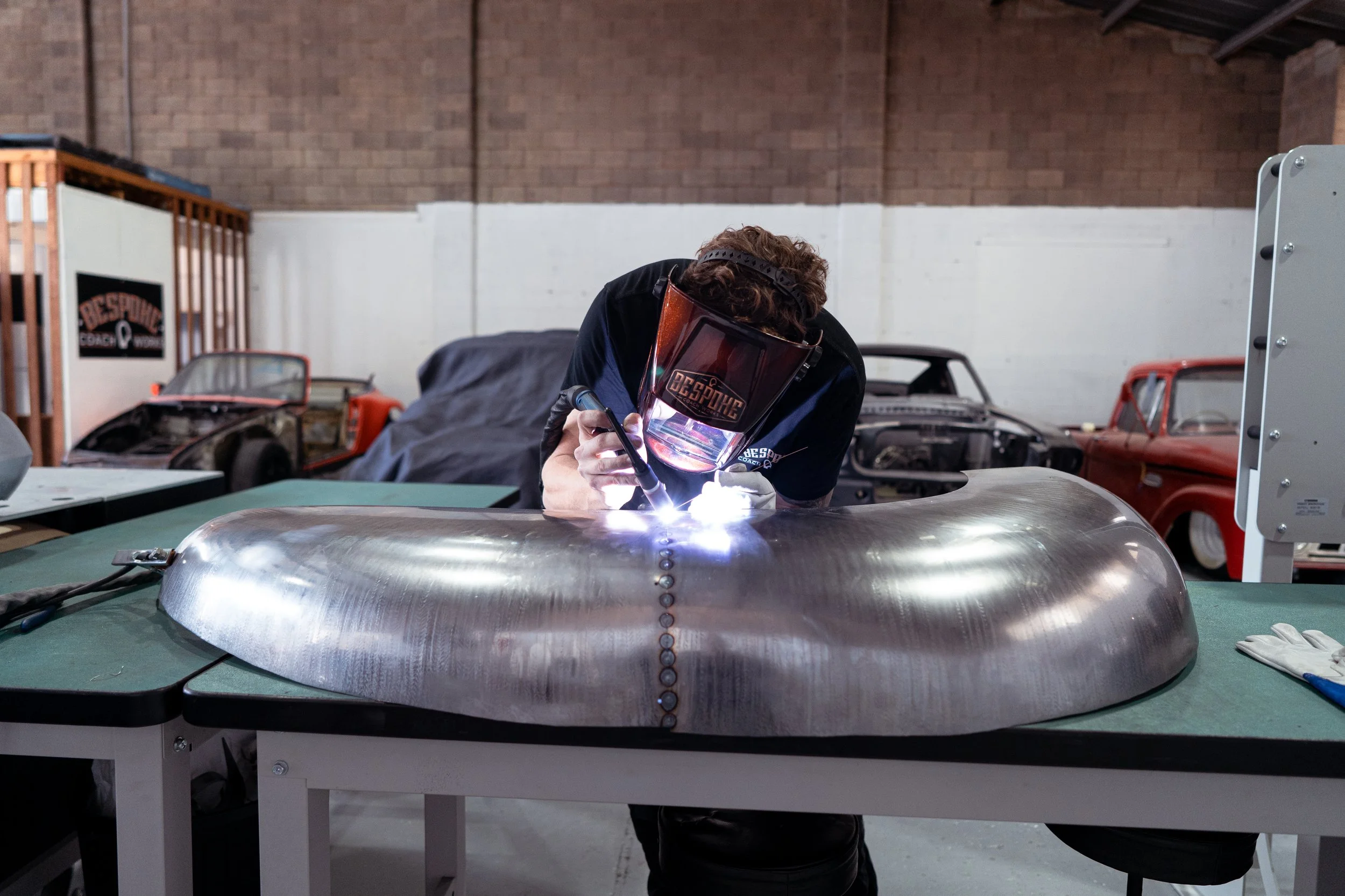 A person welding a metal automotive part in a workshop with classic cars and tools in the background.