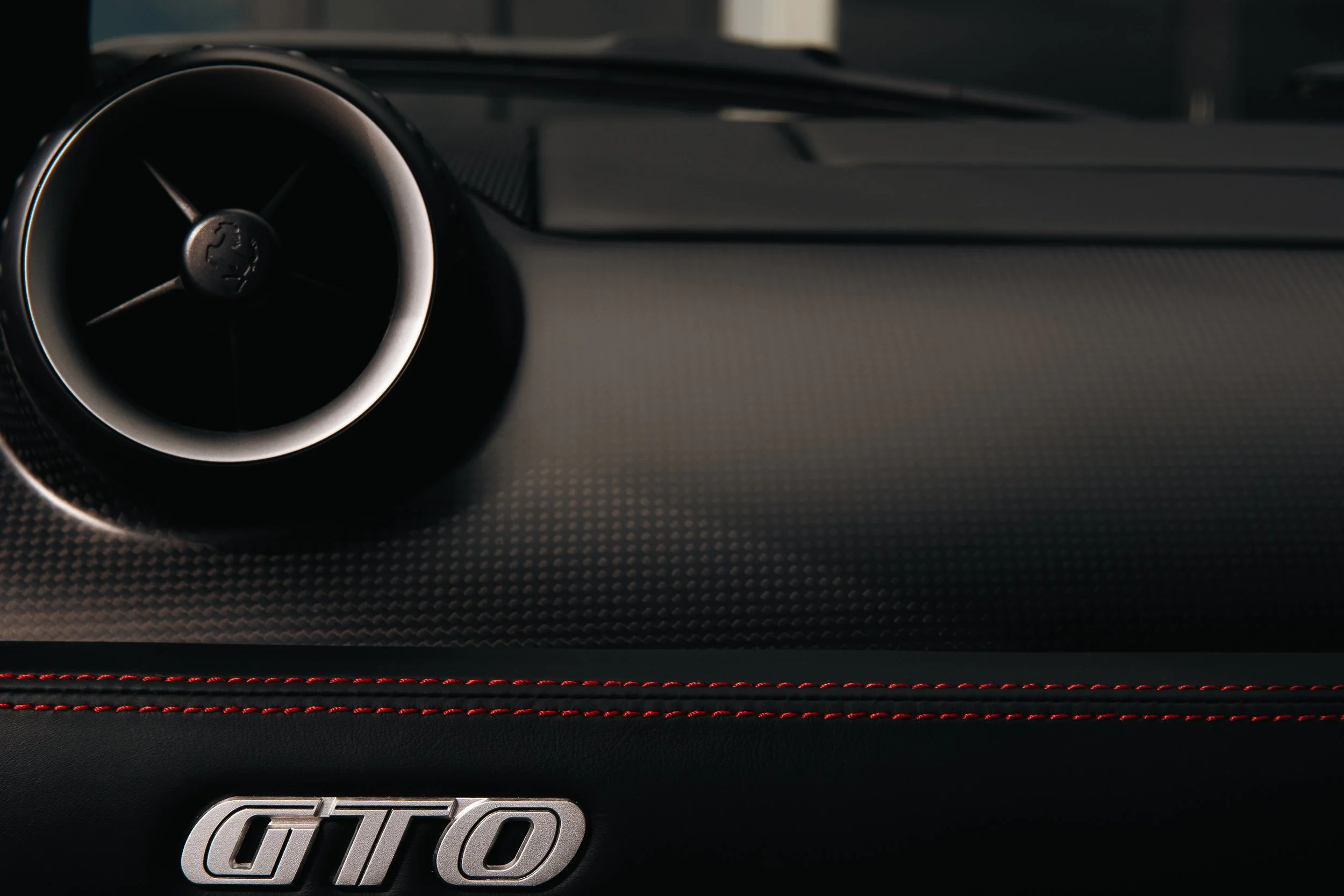 Close-up of a car dashboard featuring a round air vent with a Ferrari logo in the center and a 'GTO' emblem with red stitching below.