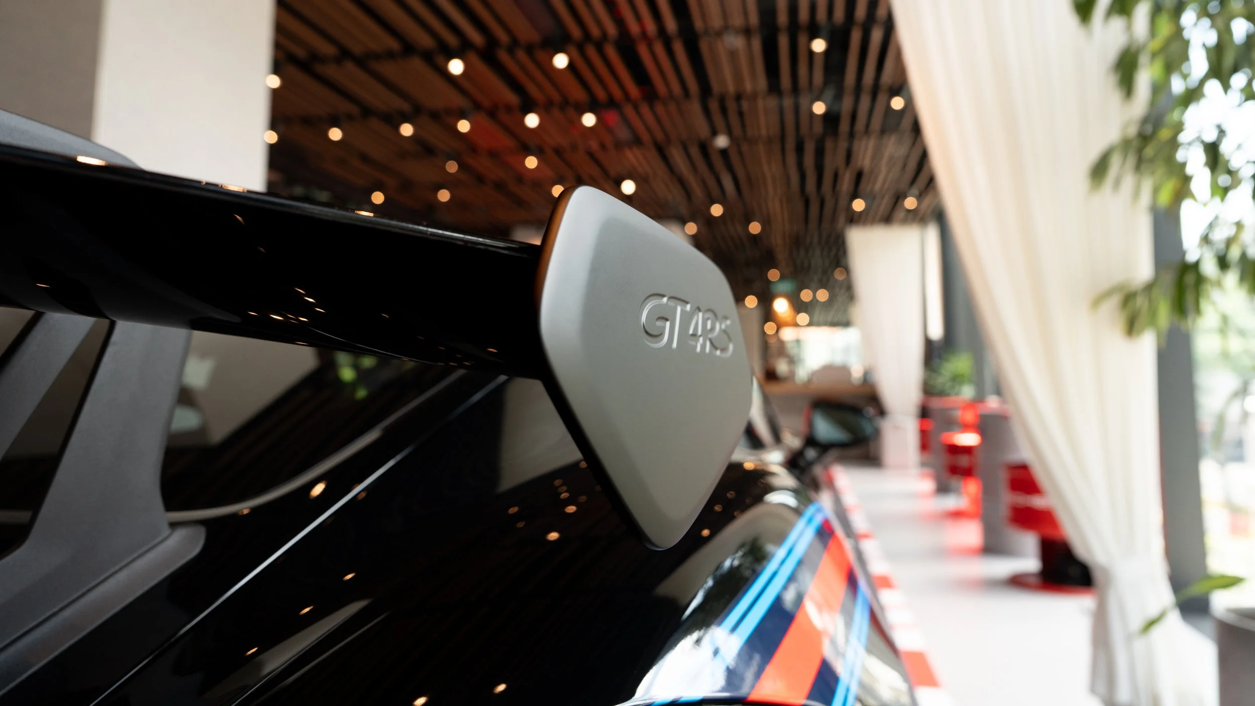 Close-up of a black Porsche with a silver GT4 RS badge, interior seen in background with curtains and potted plants.