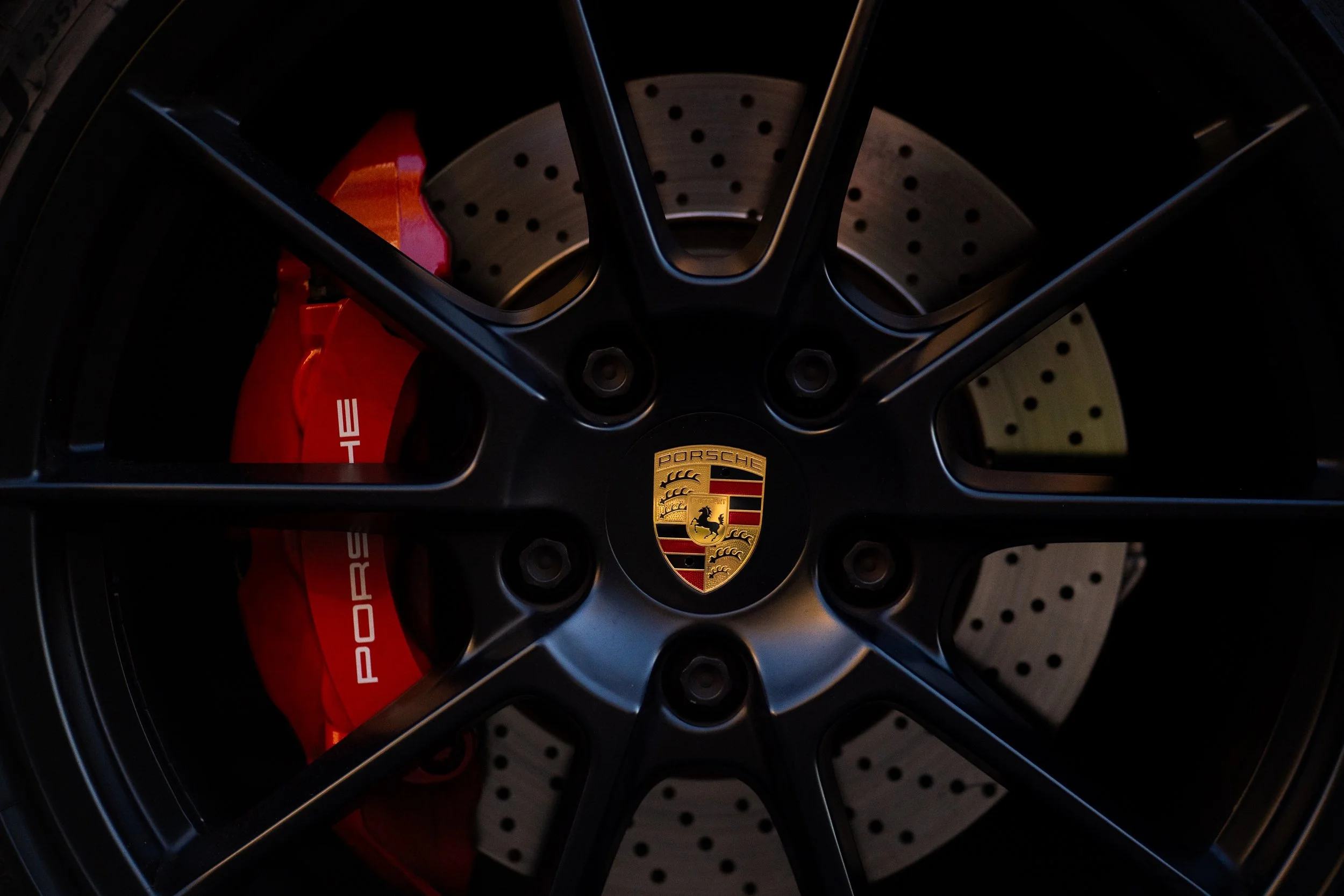 Close-up of a Porsche wheel with a black rim, red brake caliper, and perforated brake disc, featuring the Porsche emblem in the center.