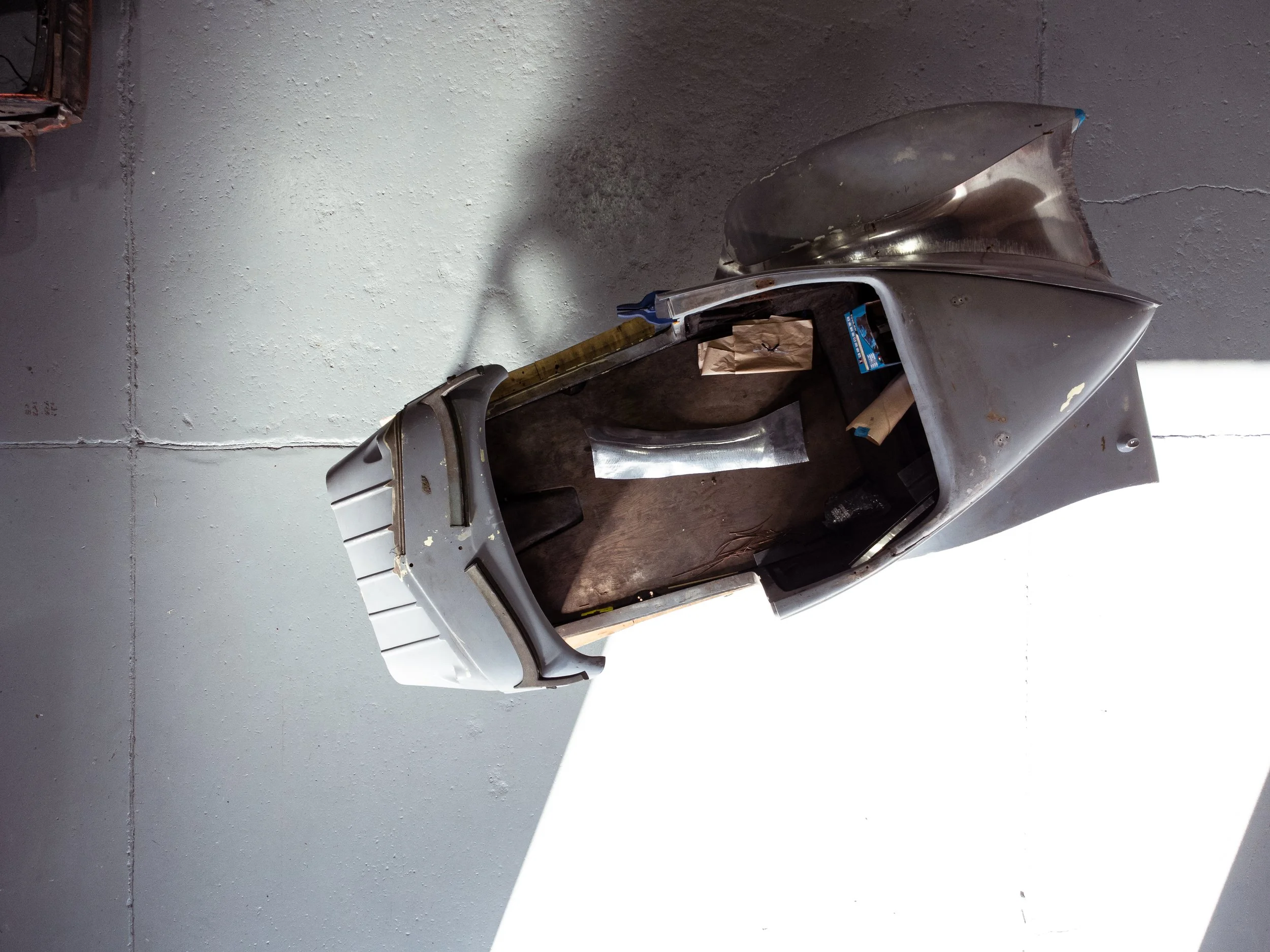 A vintage metal Vespa scooter with parts missing, placed on a wooden block against a plain gray wall.