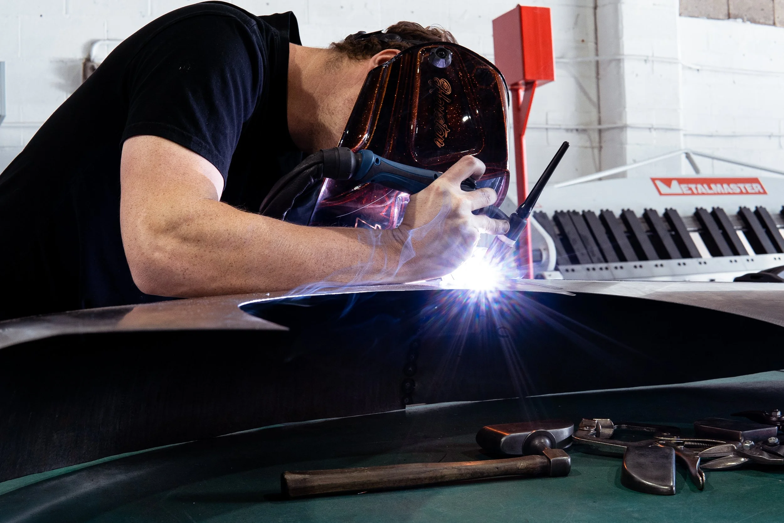 A man welding metal in a workshop, wearing a welding helmet and black shirt, with tools and equipment around him.