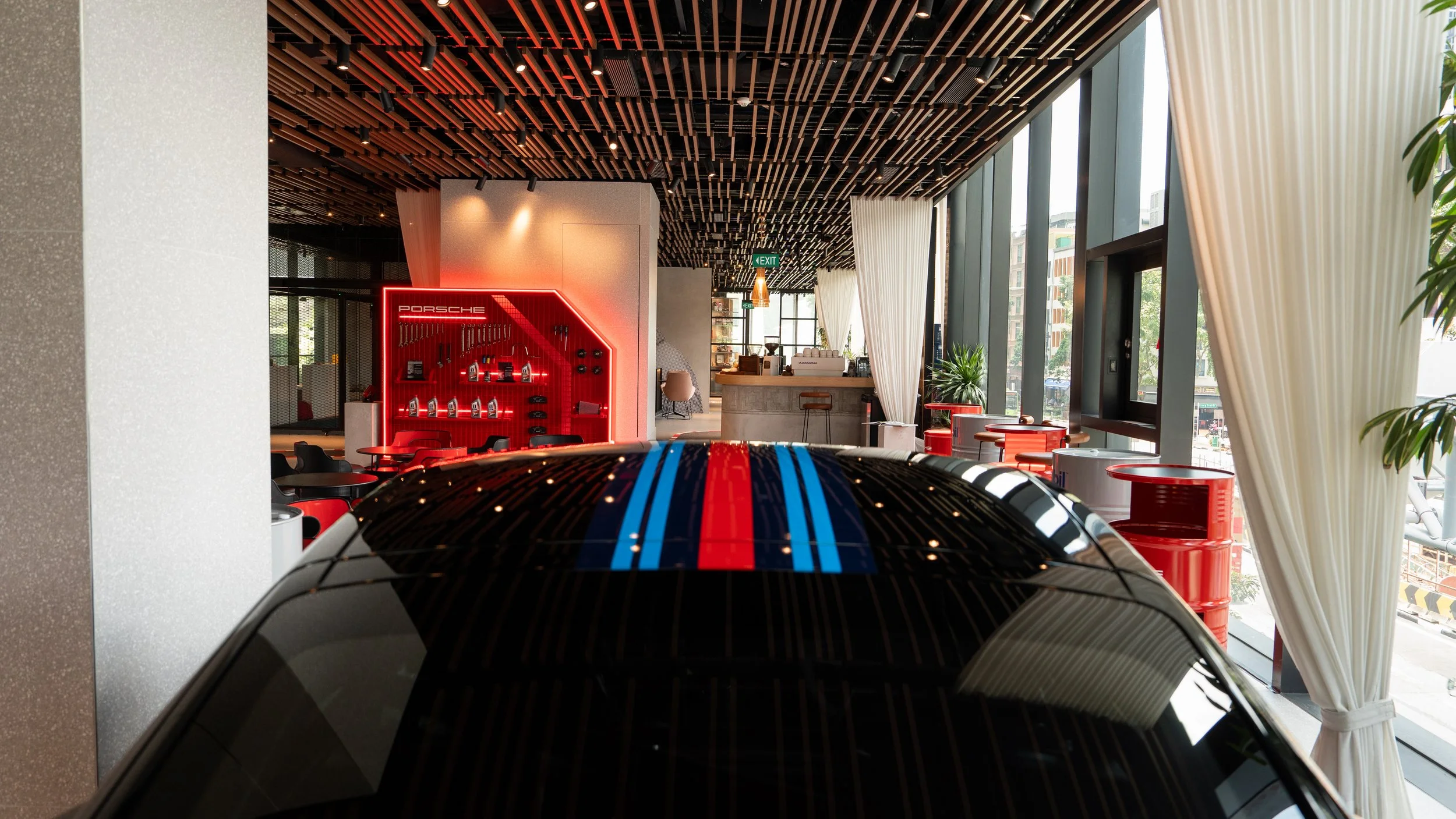 Interior of a modern car dealership with a black Porsche car in the foreground, showcasing its roof with blue and red racing stripes. The background features a Porsche display wall, seating area, large windows with white curtains, and decorative plants.
