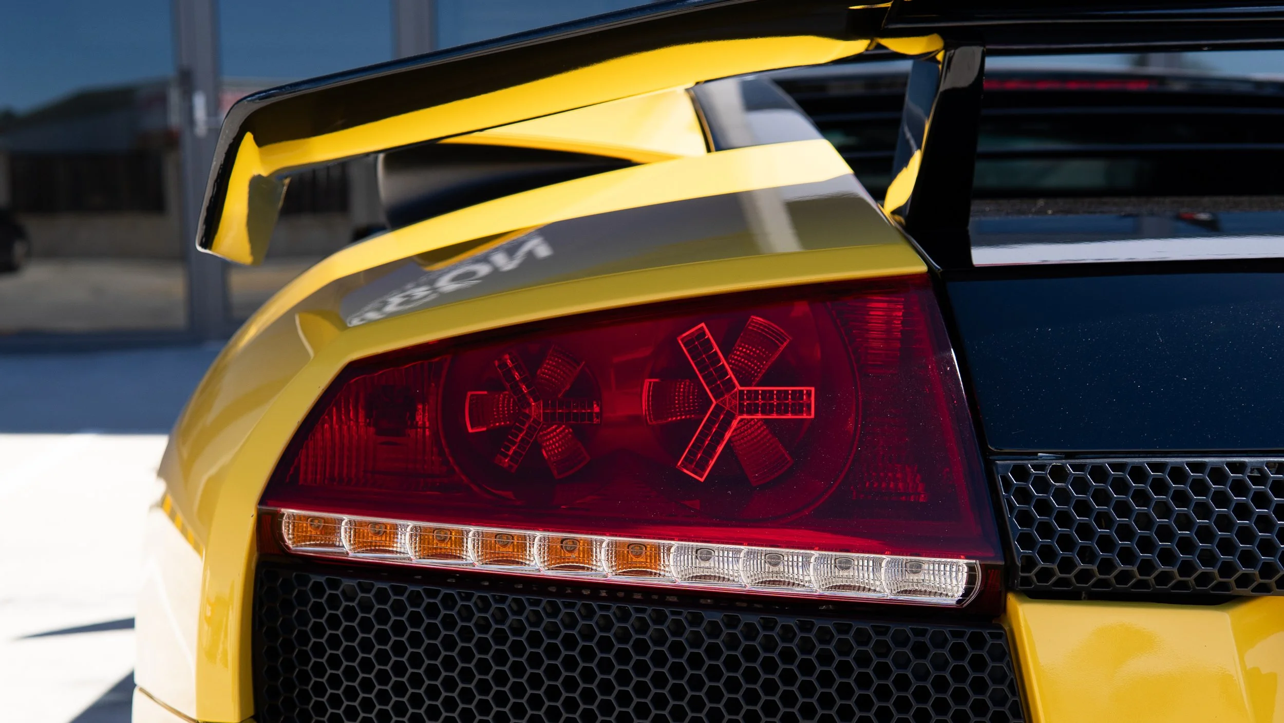 Close-up of the rear end of a yellow sports car, showing the taillight with distinctive turnover arrows and a large black rear wing spoiler.