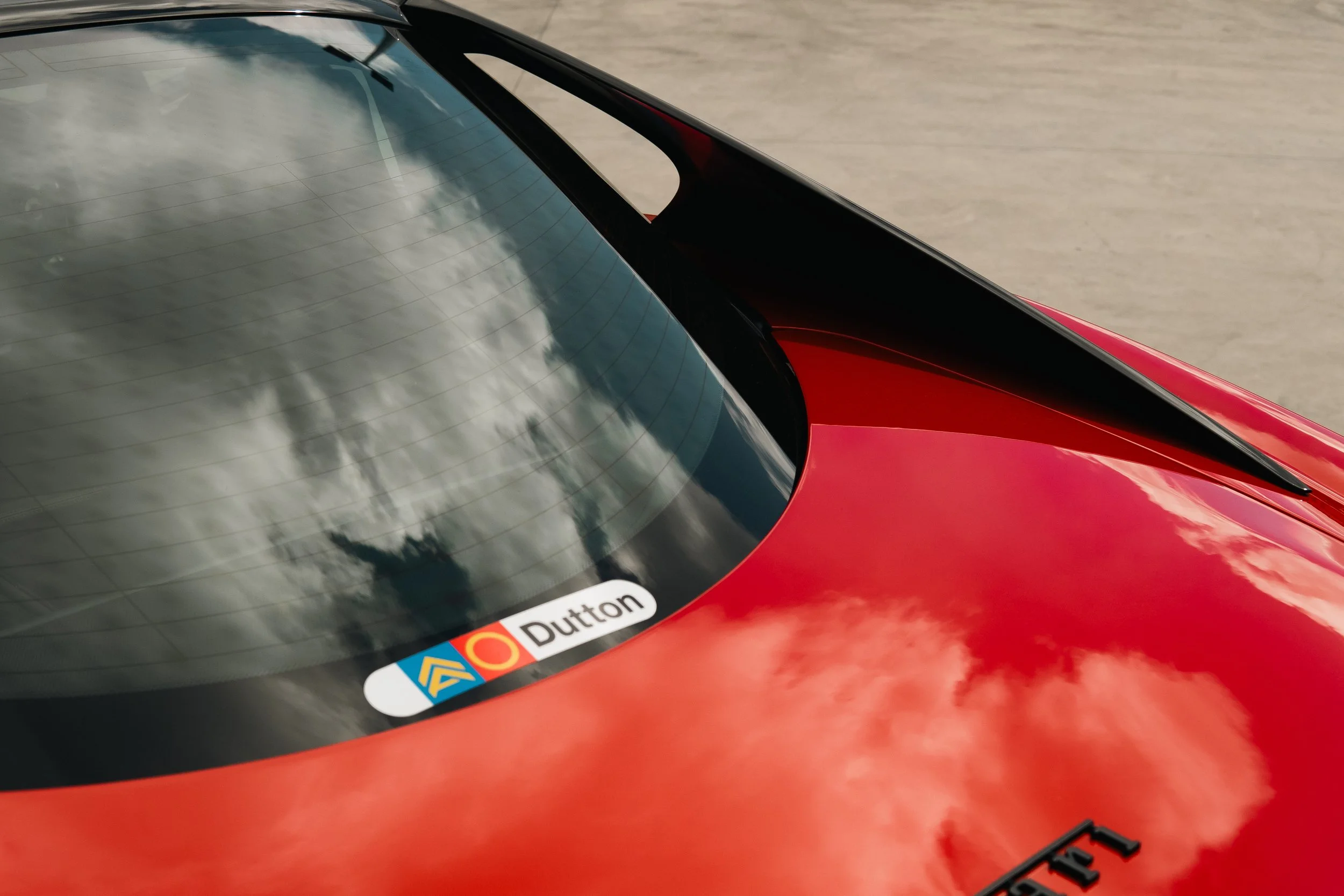Close-up of a red sports car's rear window with a sticker that reads 'Dutton' and another sticker with a logo, parked on a concrete surface.