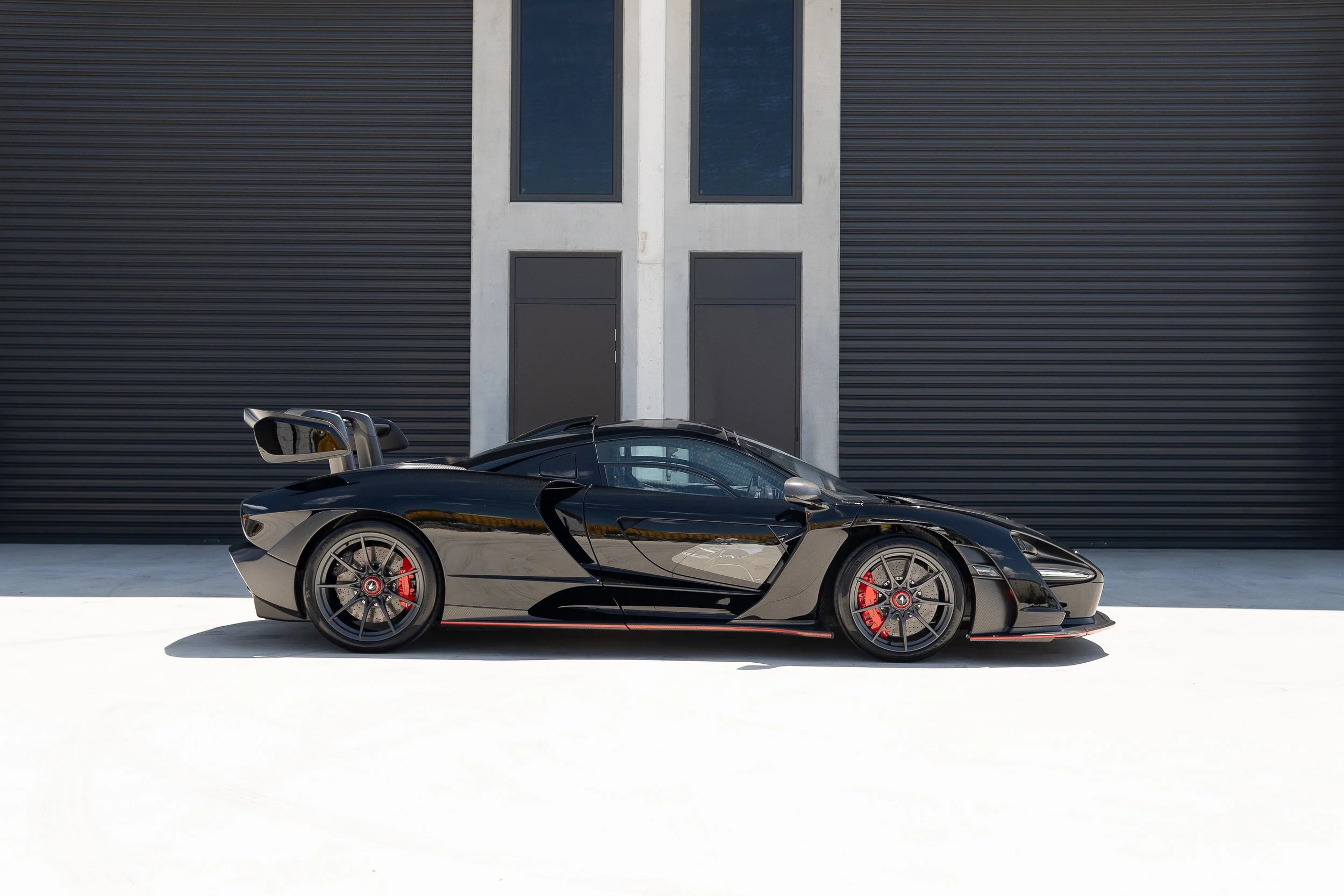 A black McLaren Senna with red brake callipers parked in front of a modern building with black and grey exterior walls and windows.
