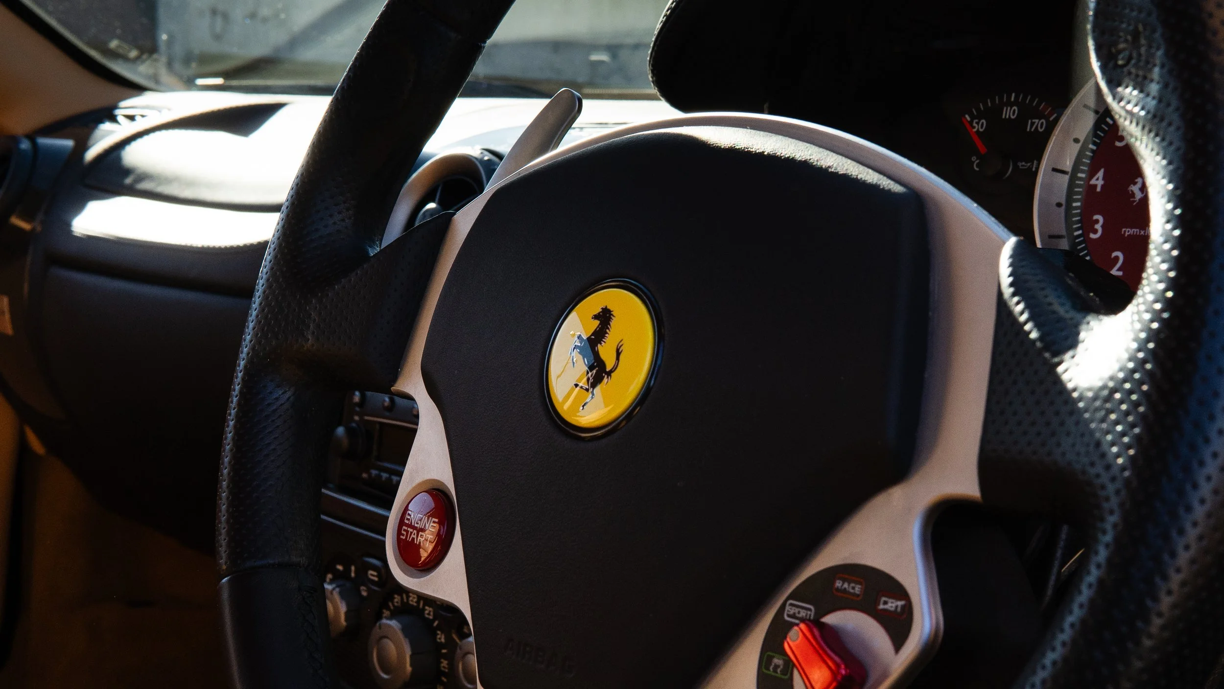Close-up of a Ferrari sports car steering wheel with a yellow prancing horse emblem center and various control buttons.