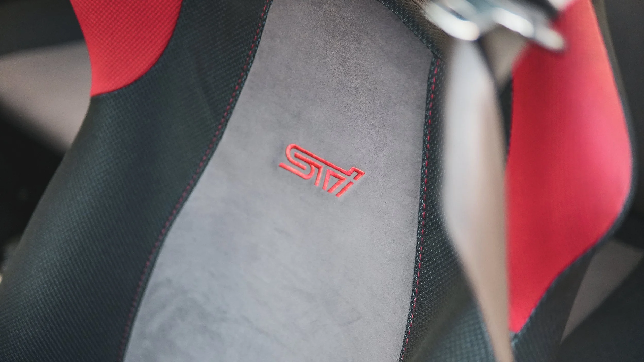 Close-up of a racing car seat with black, red, and gray fabric, featuring a red embroidered STI logo.