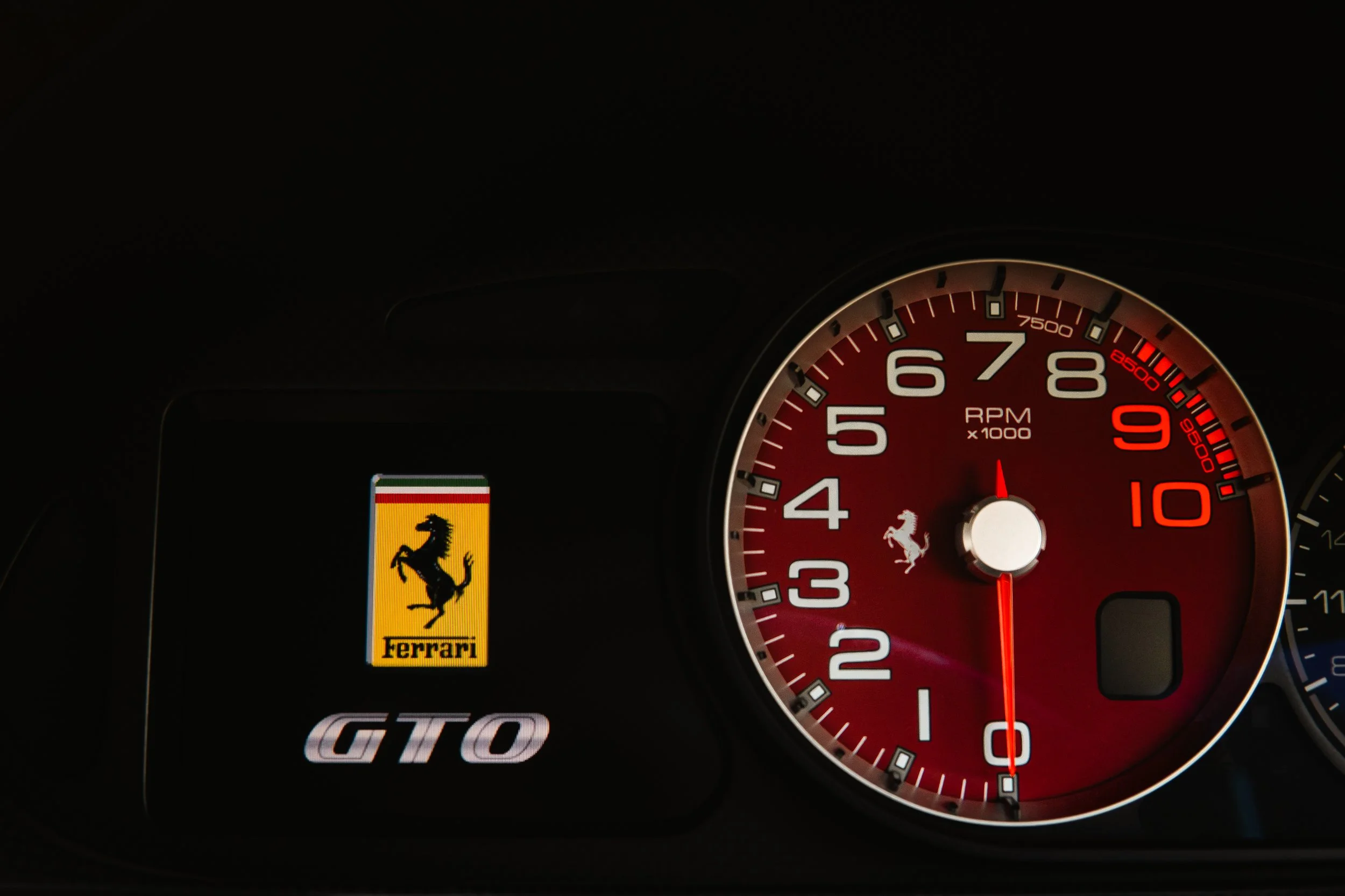 Close-up of a Ferrari dashboard showing the Ferrari logo, GT0 emblem, and a tachometer with red background and white markings.
