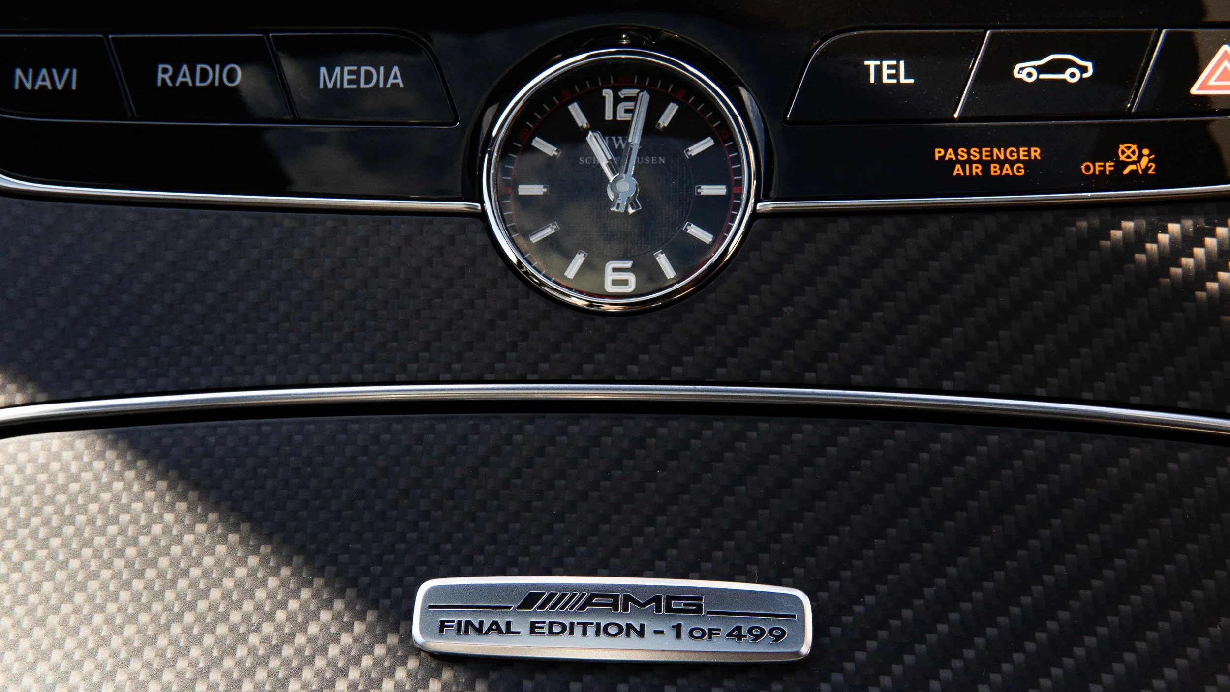 A luxury vehicle dashboard featuring an analog clock, buttons labeled NAVI, RADIO, MEDIA, TEL, an indicator for passenger airbag, and a badge that reads 'AMG FINAL EDITION - 1 OF 499' with a carbon fiber textured background.