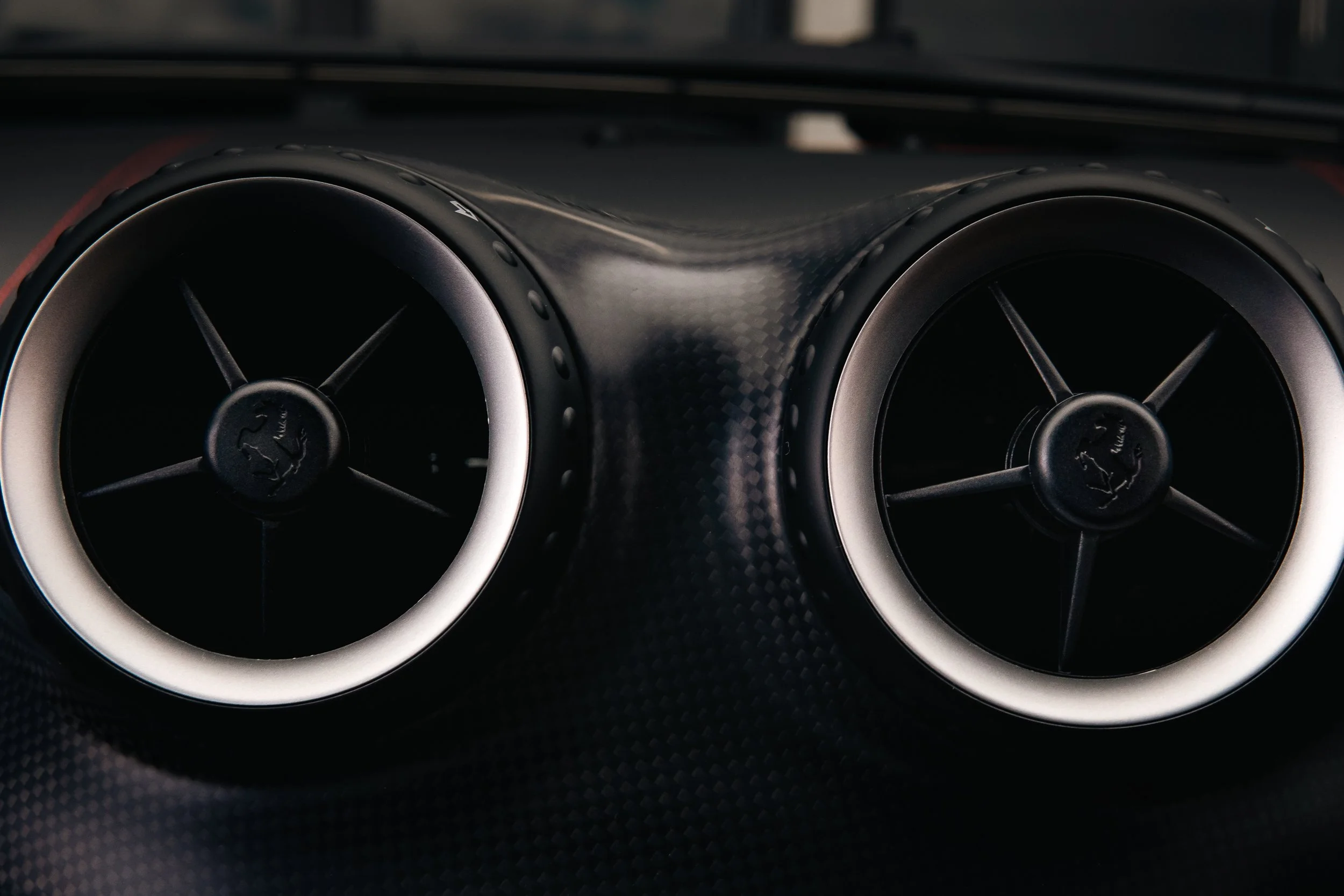 Two circular black car air vents with silver trim, mounted on a textured dashboard of a Ferrari.