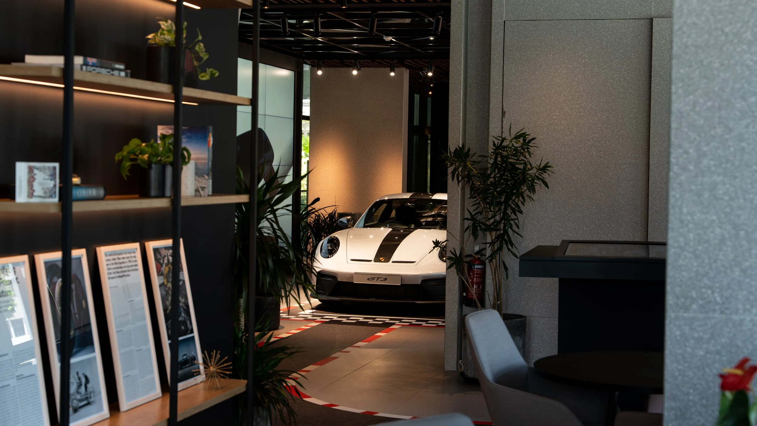 A white Porsche 911 GT3 sports car with black accents displayed indoors, framed by plants and modern interior decor.